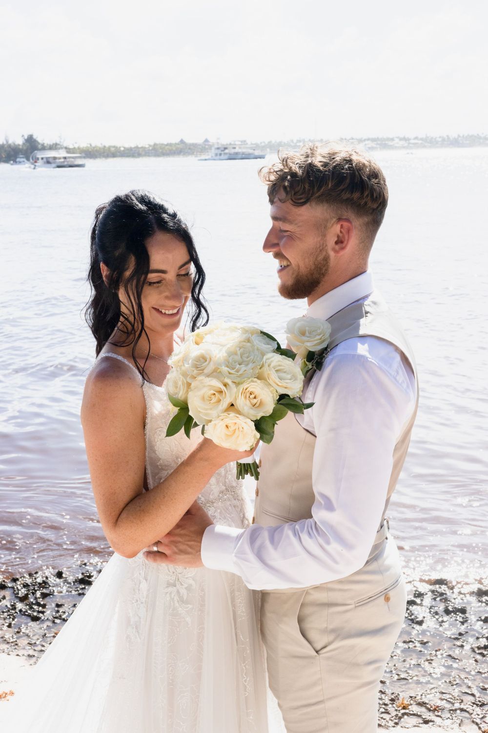 A newlywed couple embraces by the water holding a bouquet of white roses.