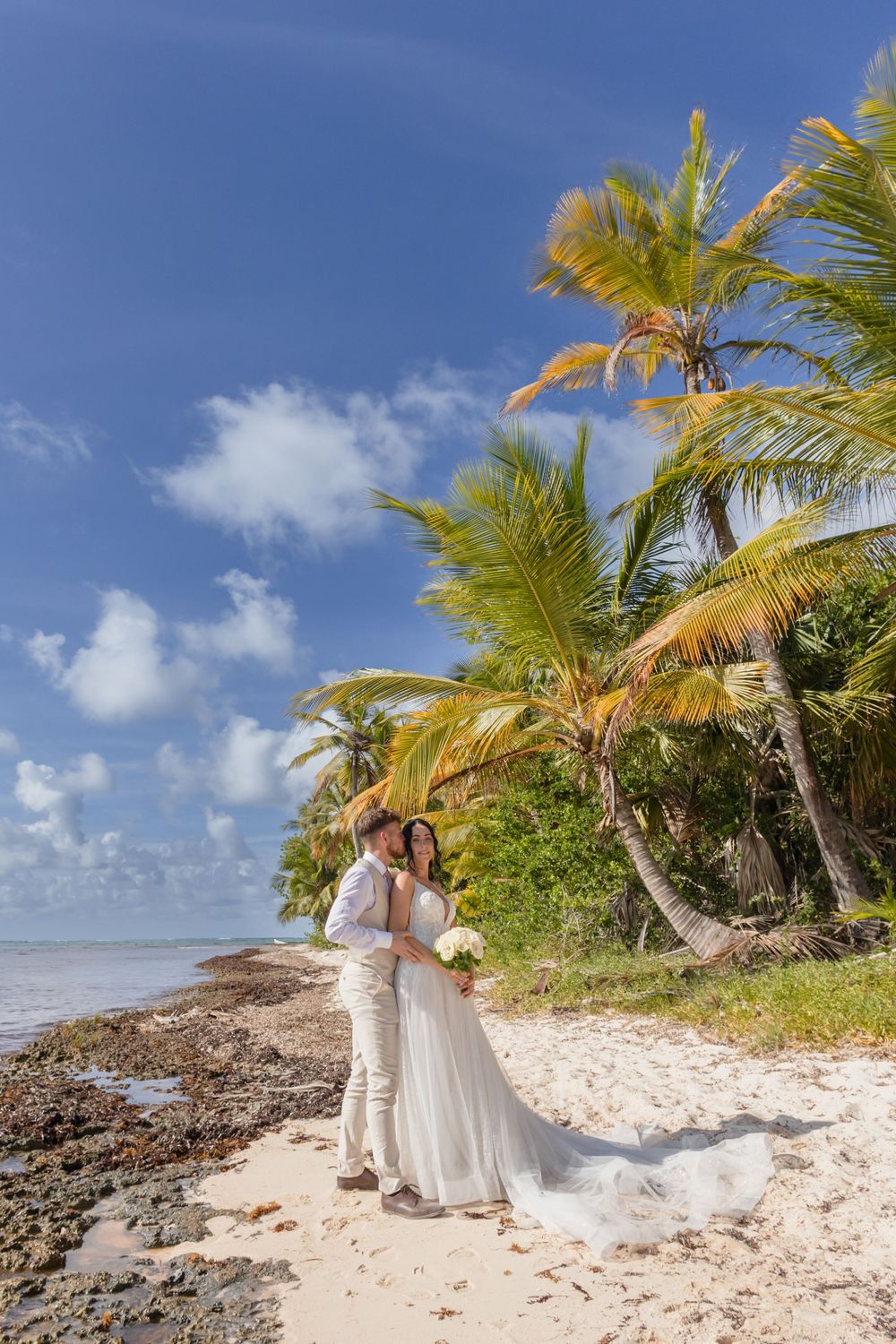 A couple in white attire poses on a scenic beach with palm trees and rocky coastline in the background.