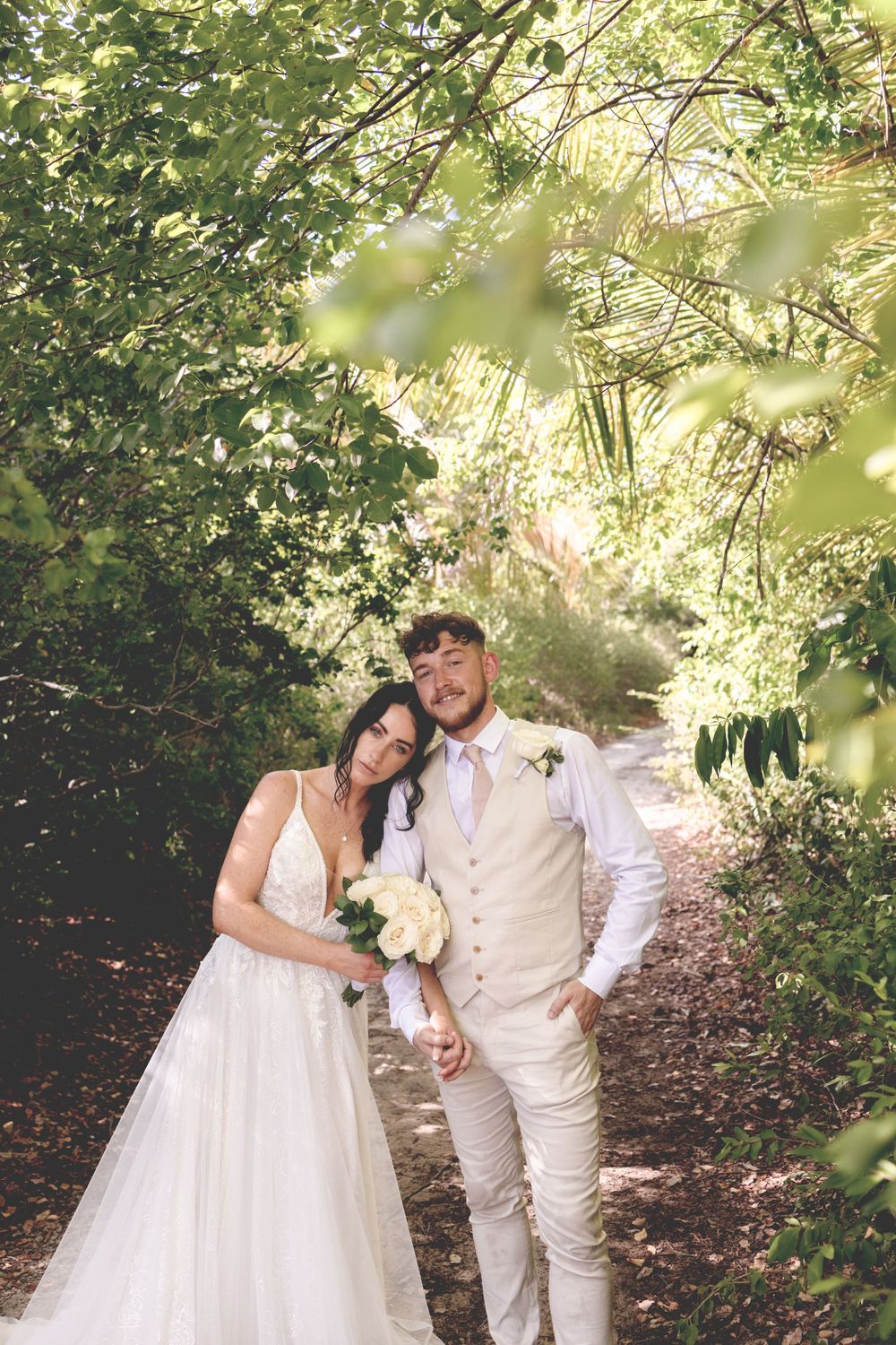 A romantic outdoor wedding scene with a couple in white and beige attire embracing on a wooded path.