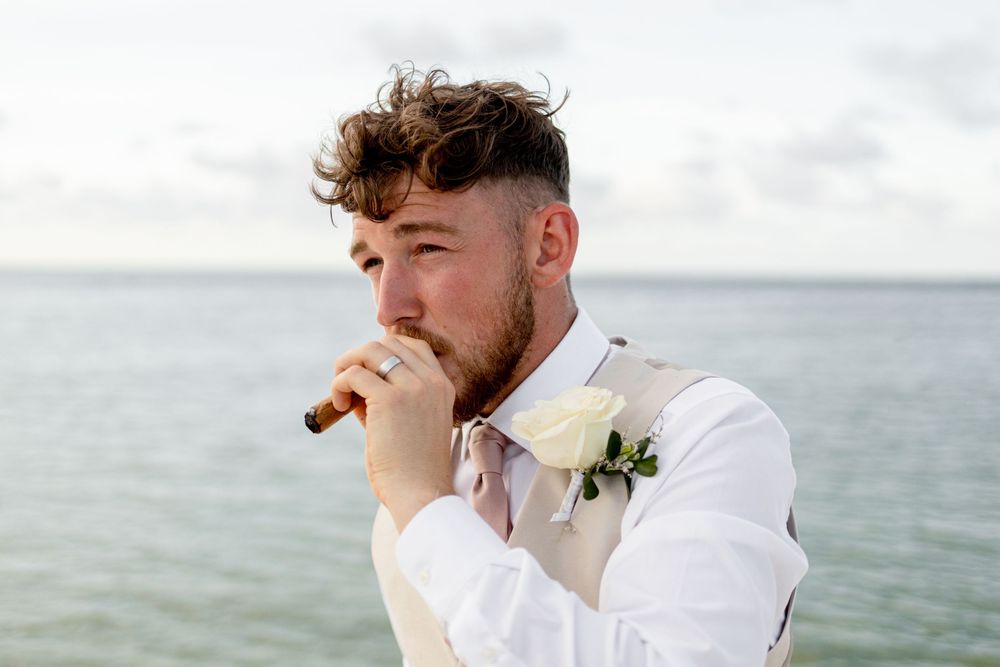A groom in a white shirt and boutonniere smokes a cigar while looking out over the ocean on his wedding day.