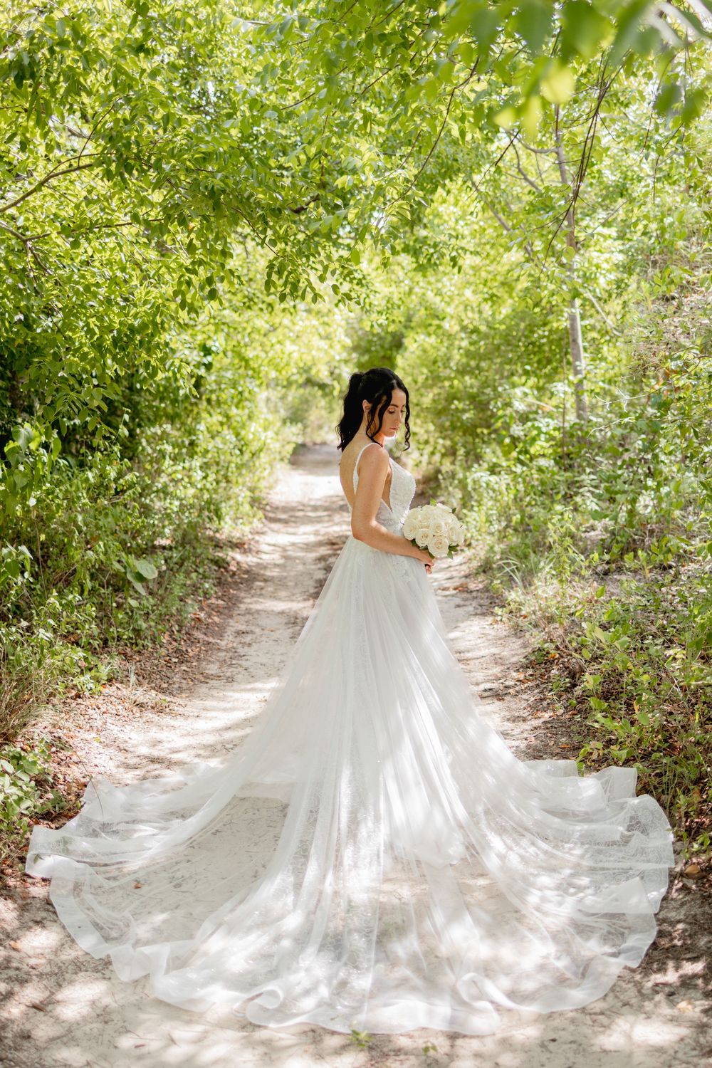 A flowing white wedding dress with a long train trails through a sunlit garden path surrounded by lush greenery.