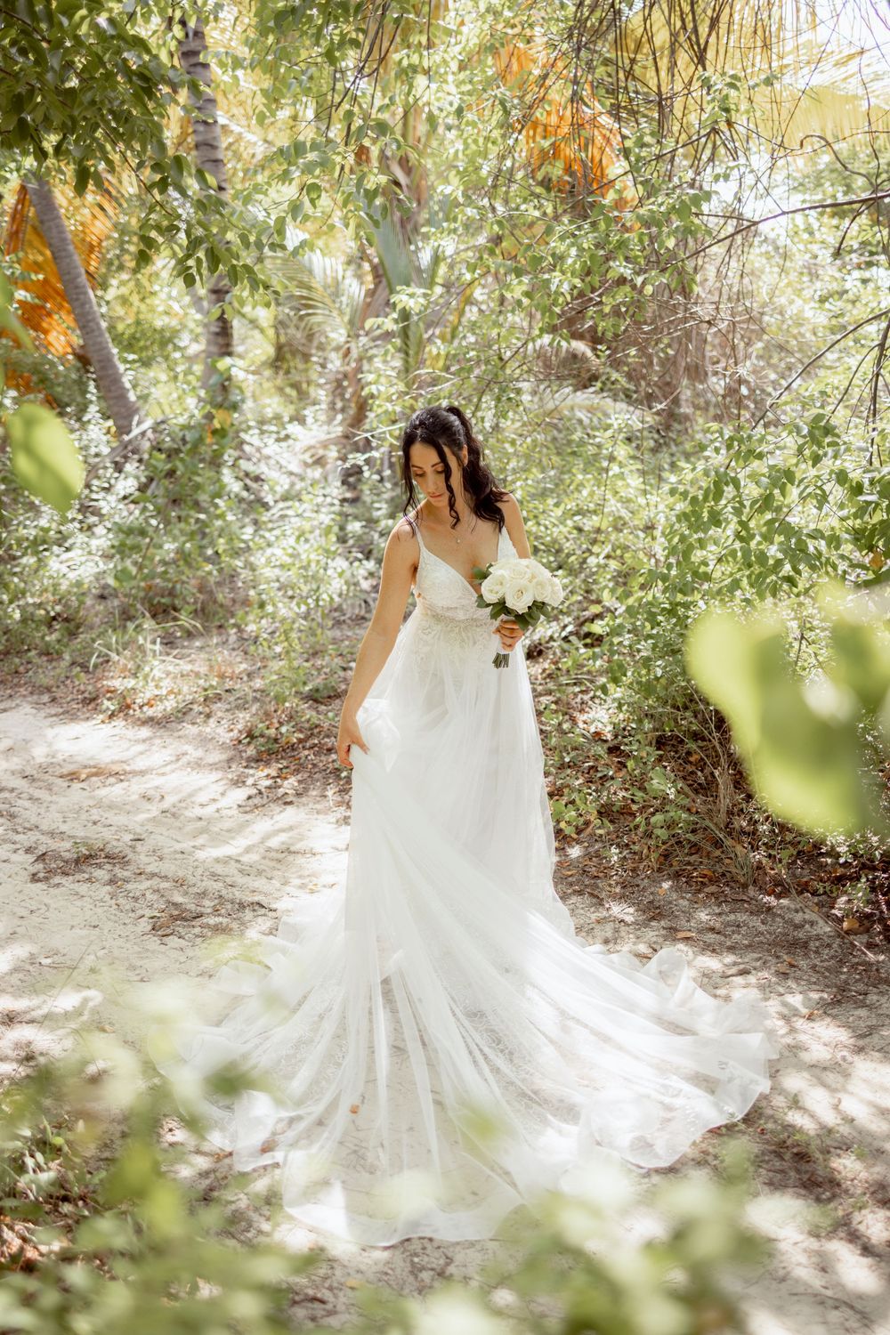A flowing white wedding dress cascades across a sunlit forest path surrounded by lush greenery and dappled light.