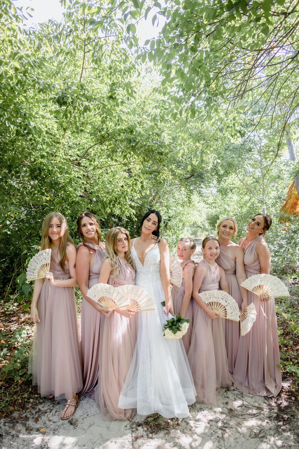 Wedding party in blush pink bridesmaid dresses poses together under trees in an outdoor garden setting.