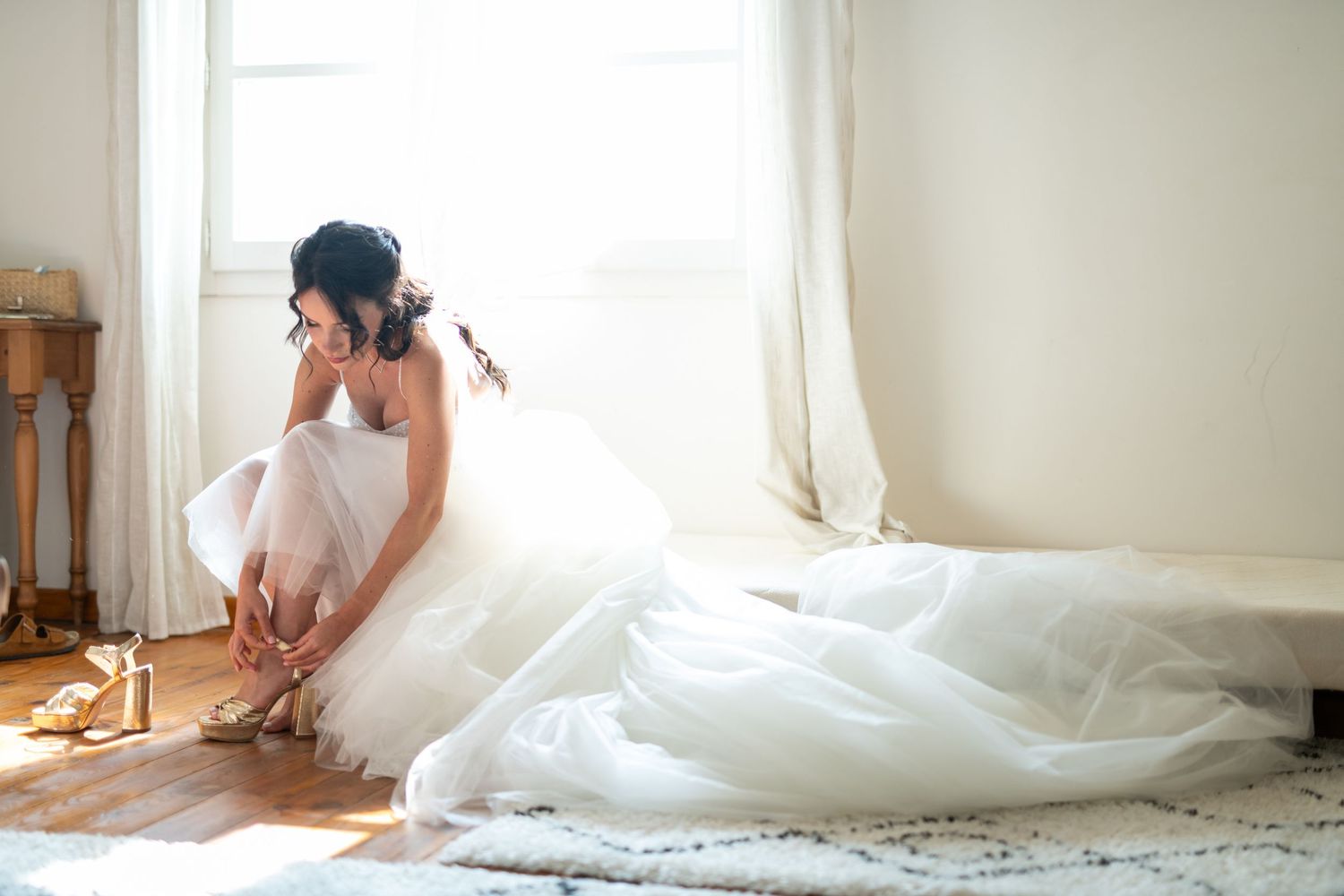 Mariée assise près d’une fenêtre, robe blanche étalée, lumière naturelle douce et moment intime des préparatifs.