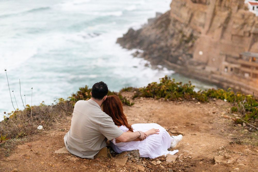 Two people sit together on a rocky cliff overlooking the ocean at sunset.