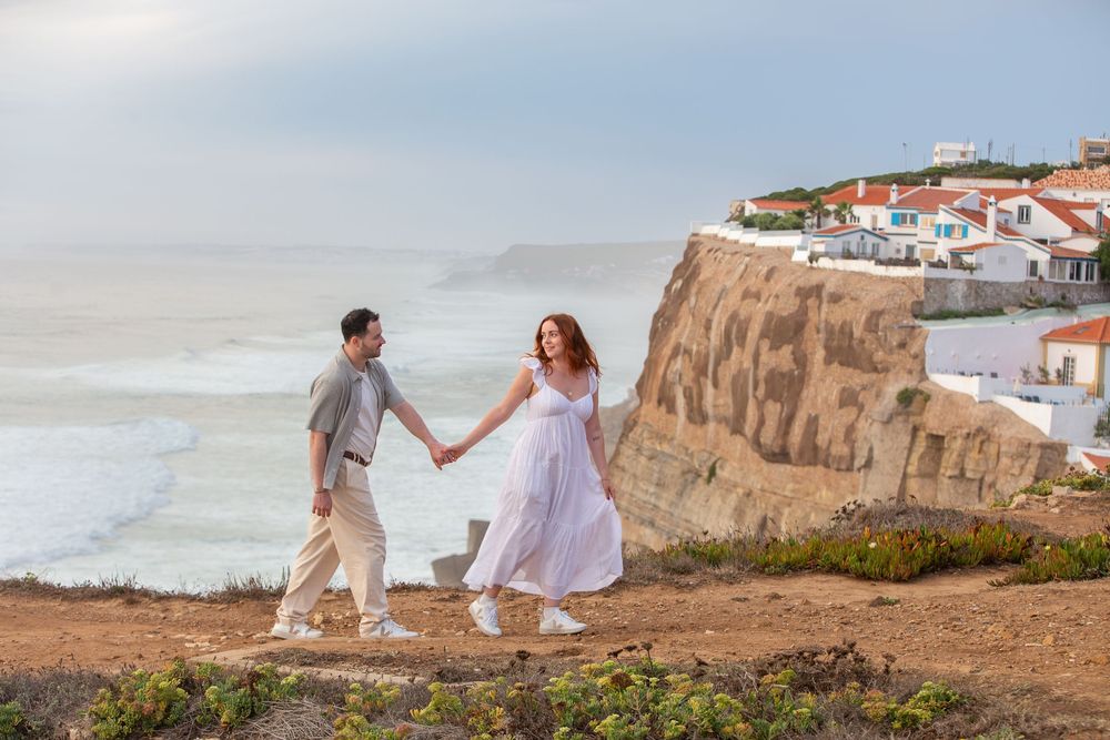 A couple in white outfits walks along a scenic coastal cliff path with ocean views and white buildings in the distance.