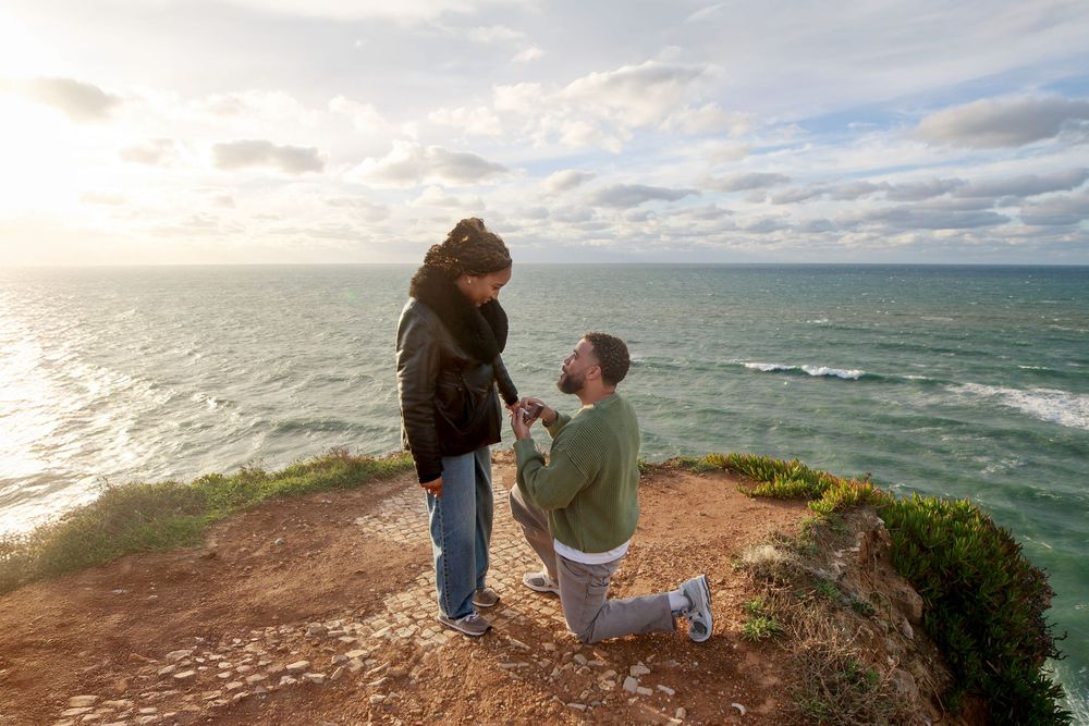A person sits on a rocky coastal cliff overlooking the turquoise ocean waters and dramatic cloudy sky.