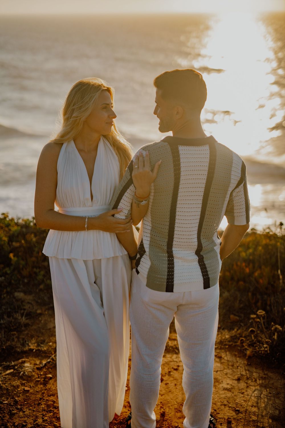 A couple in white outfits embraces during golden hour sunset on a coastal cliff overlooking the ocean.