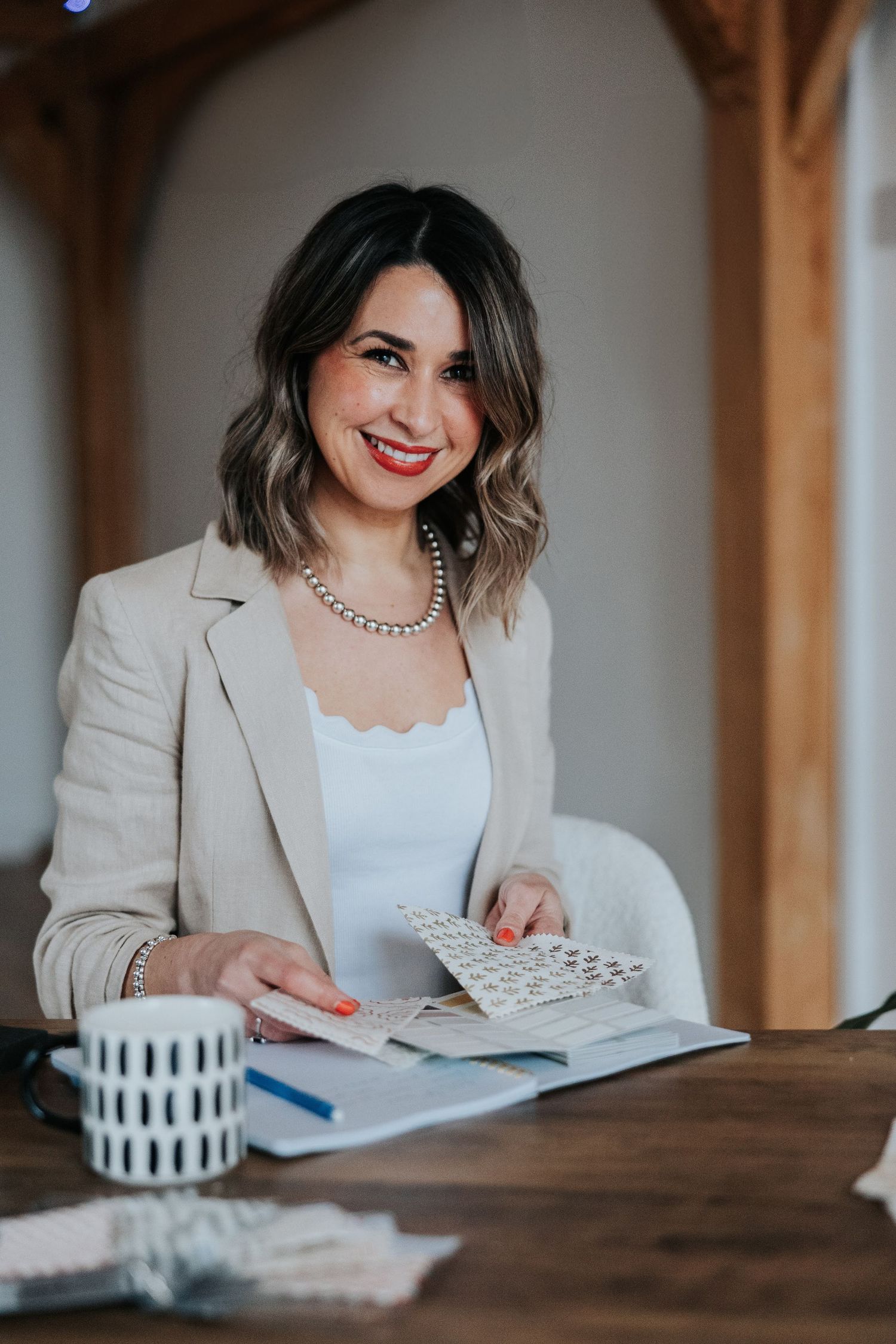 A person in a beige blazer writes in a notebook at a wooden table with a striped mug nearby.