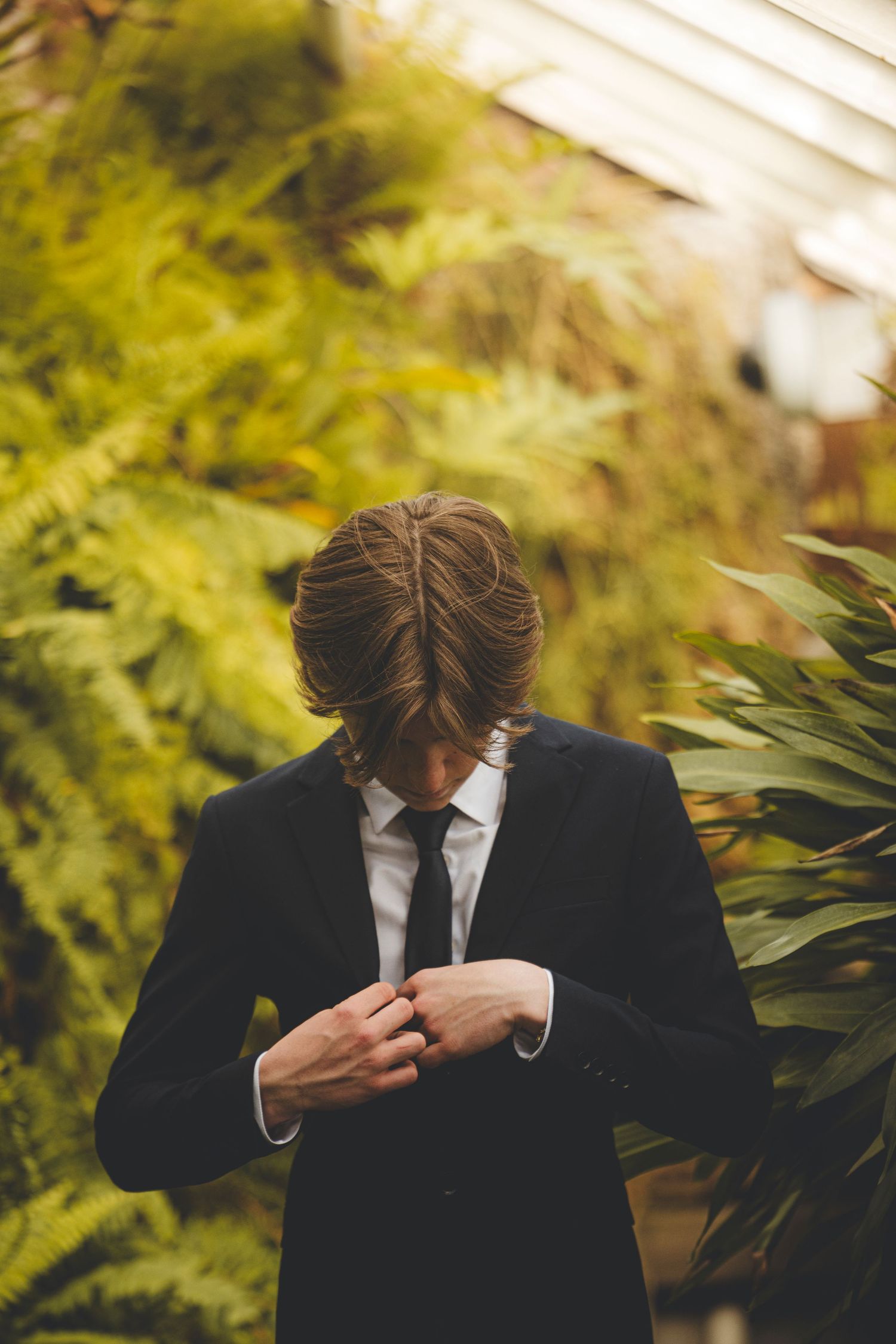Figure in dark suit adjusts tie while standing among lush green foliage.