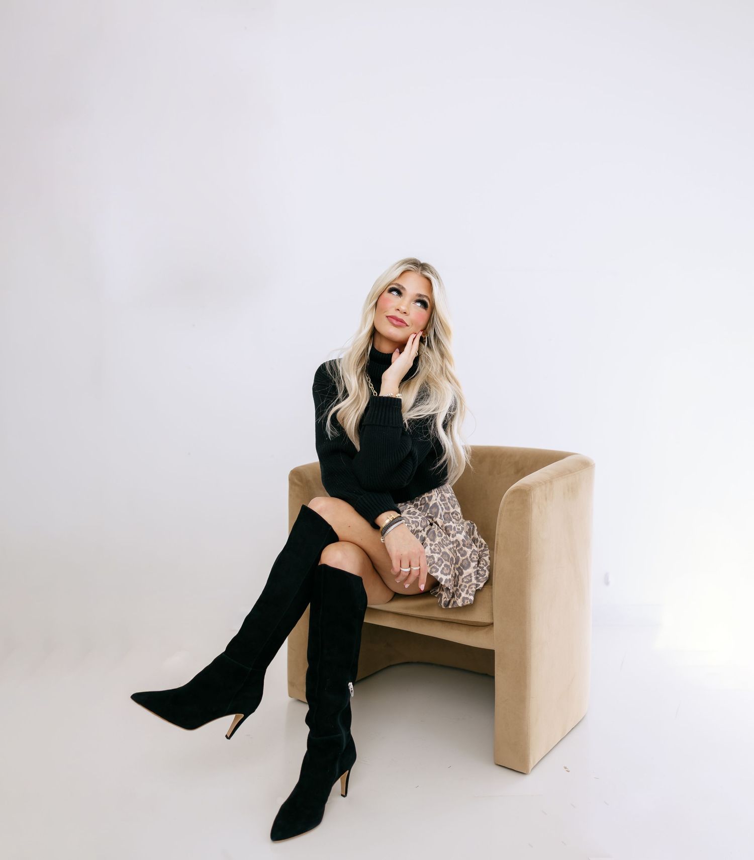 Woman in black sweater and tall boots poses on a tan upholstered chair against a white studio backdrop.