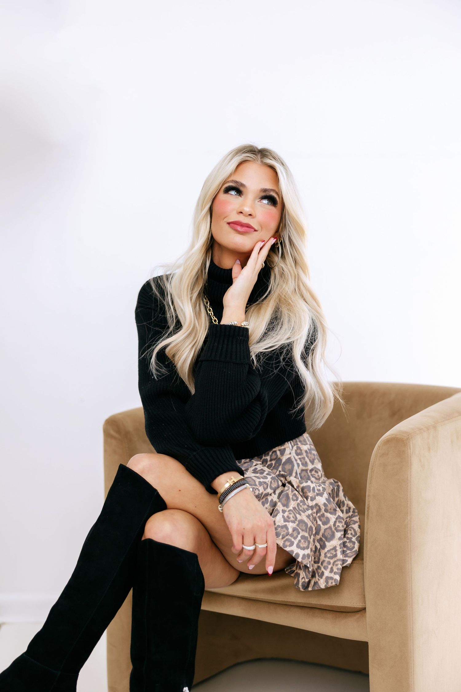 A person in a black sweater and snakeskin skirt sits thoughtfully in a tan chair against a white backdrop.