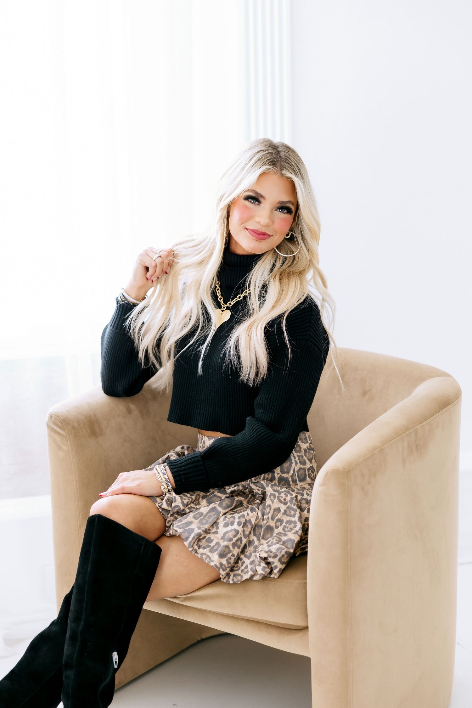 Stylish winter outfit featuring a black turtleneck sweater, leopard print skirt and tall black boots on beige accent chair.
