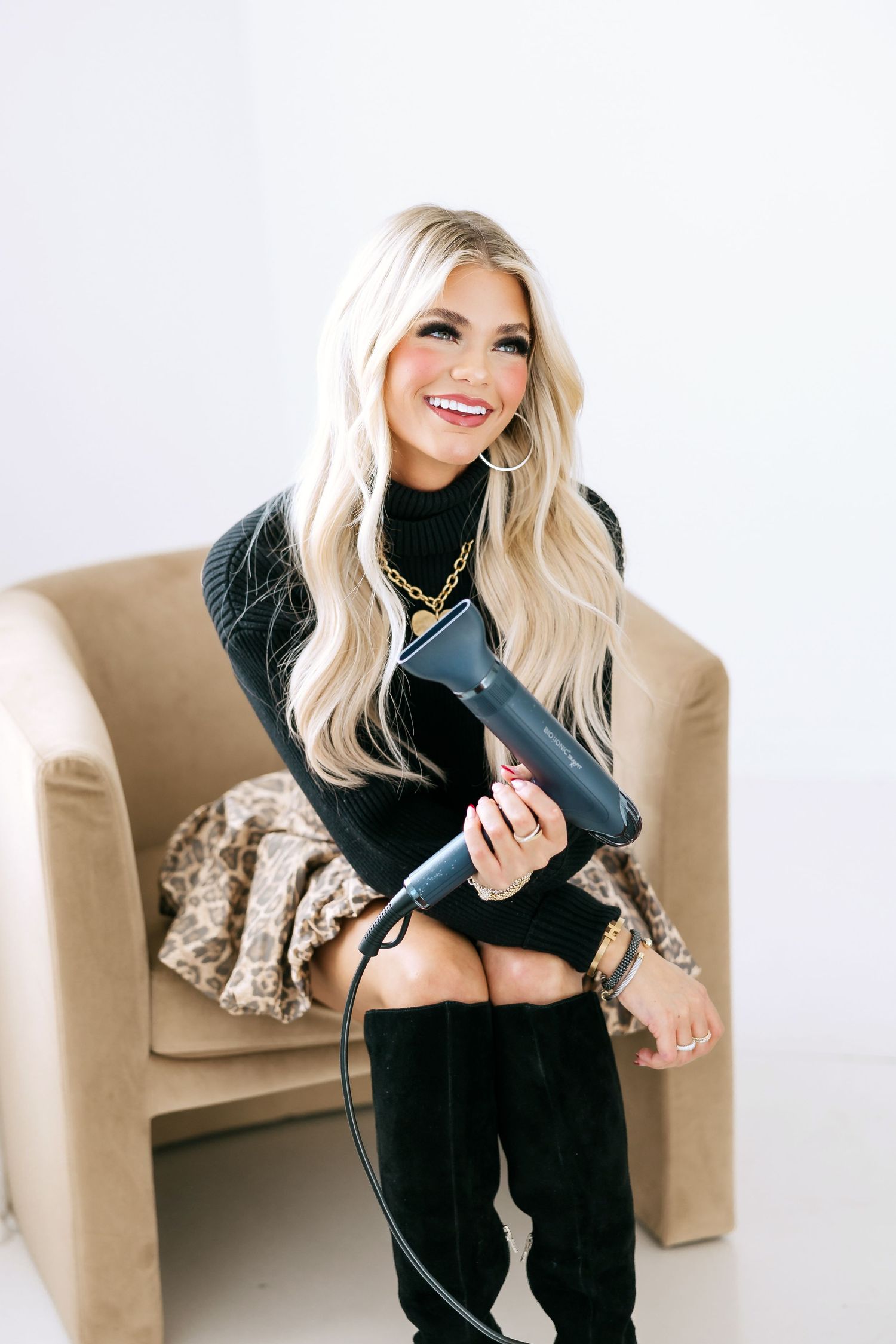 Professional stylist with long blonde hair wearing black sweater holds hair dryer while sitting in tan armchair.