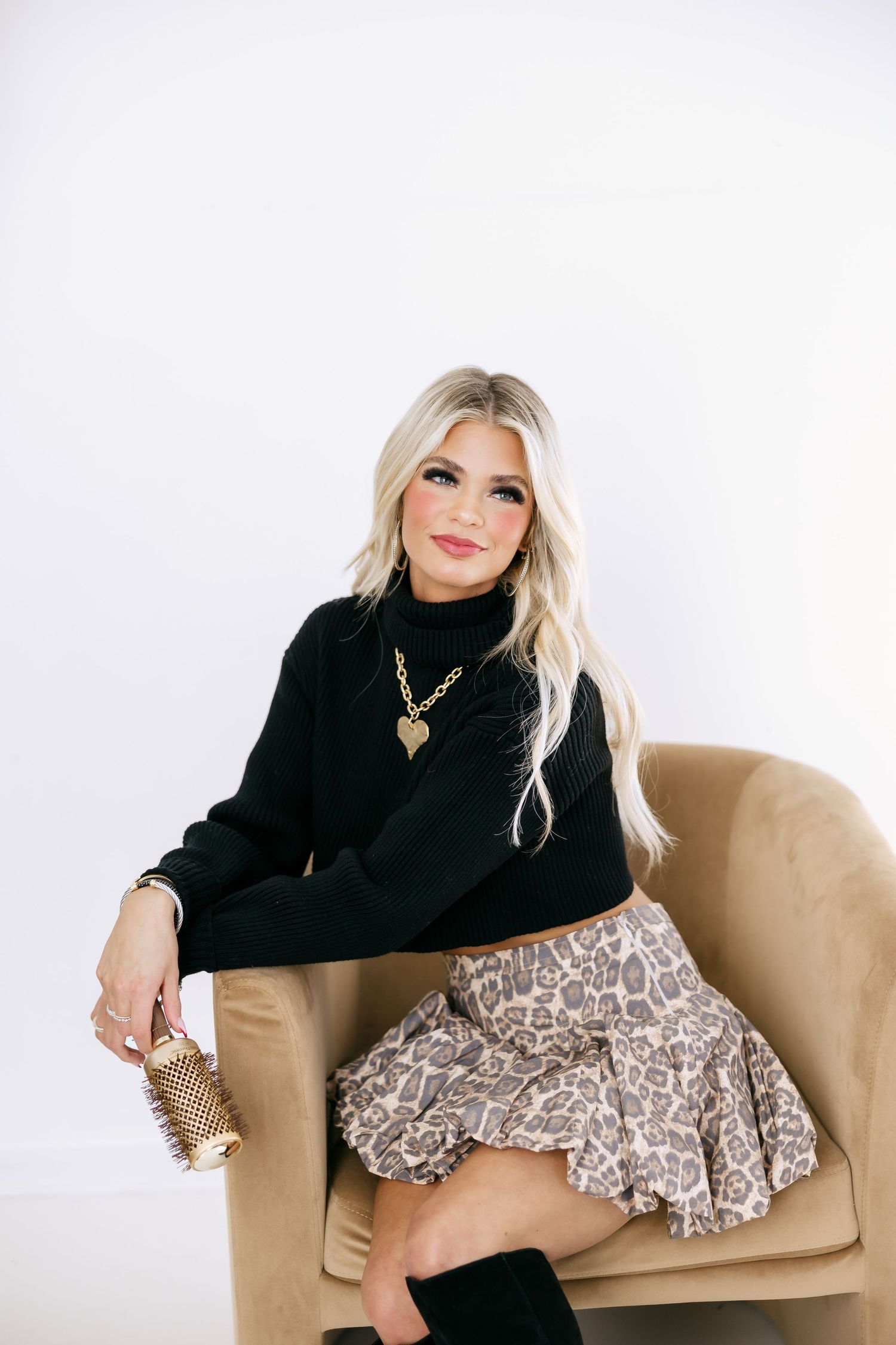 Fashion model in black sweater and patterned skirt poses elegantly on tan armchair against white background.