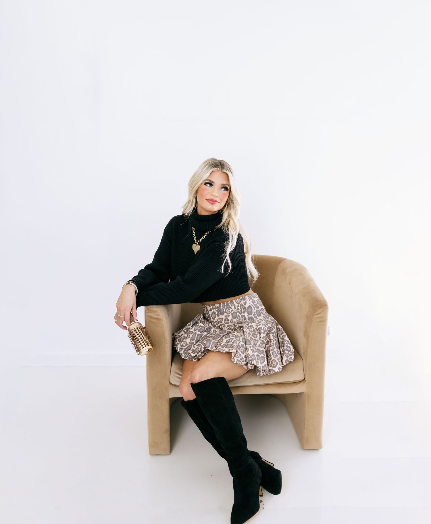 Fashion model in black sweater and patterned skirt poses on tan armchair with tall black boots against white background.
