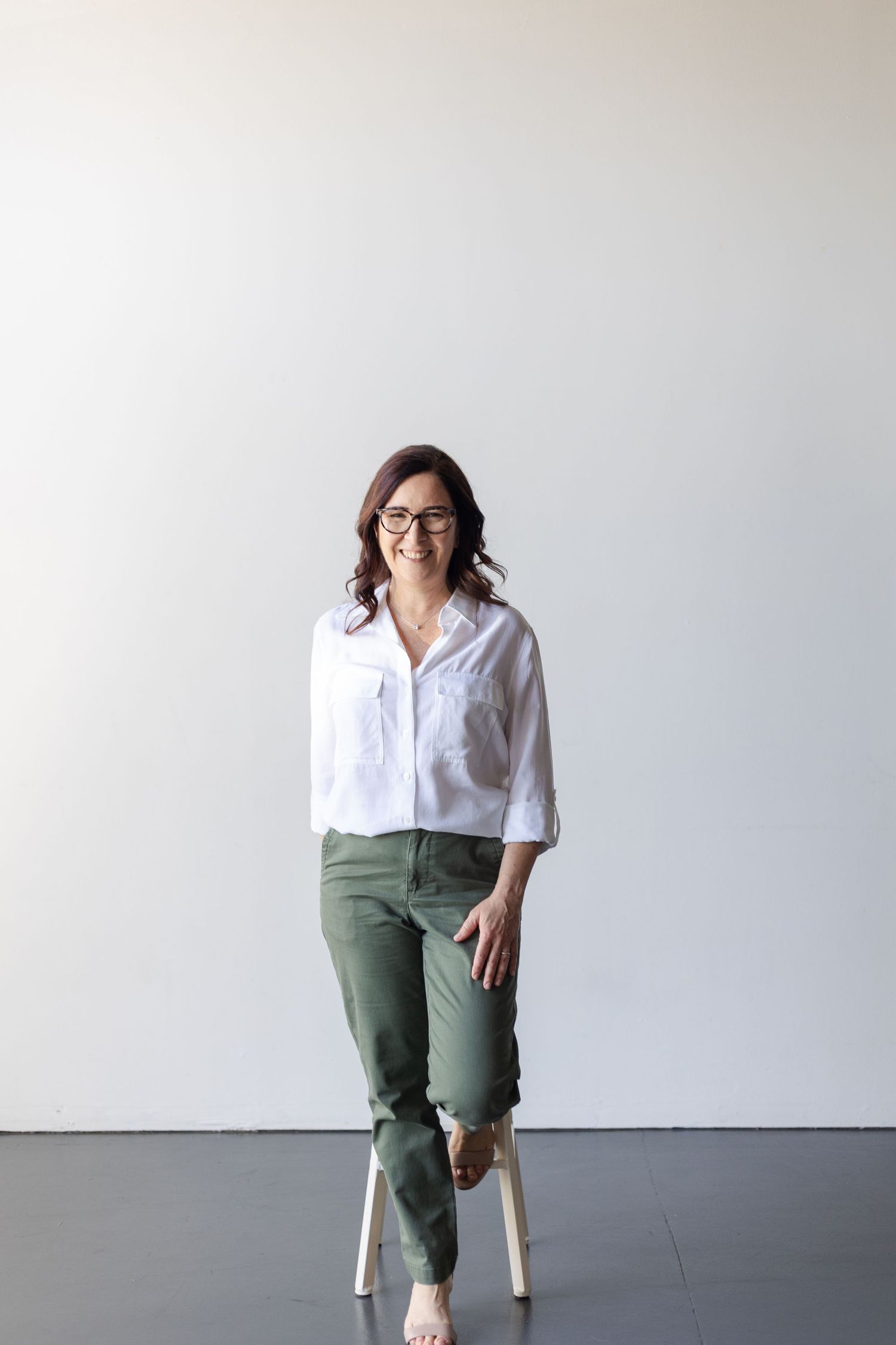 Casual portrait of woman in white shirt and olive pants sitting on stool against white wall.