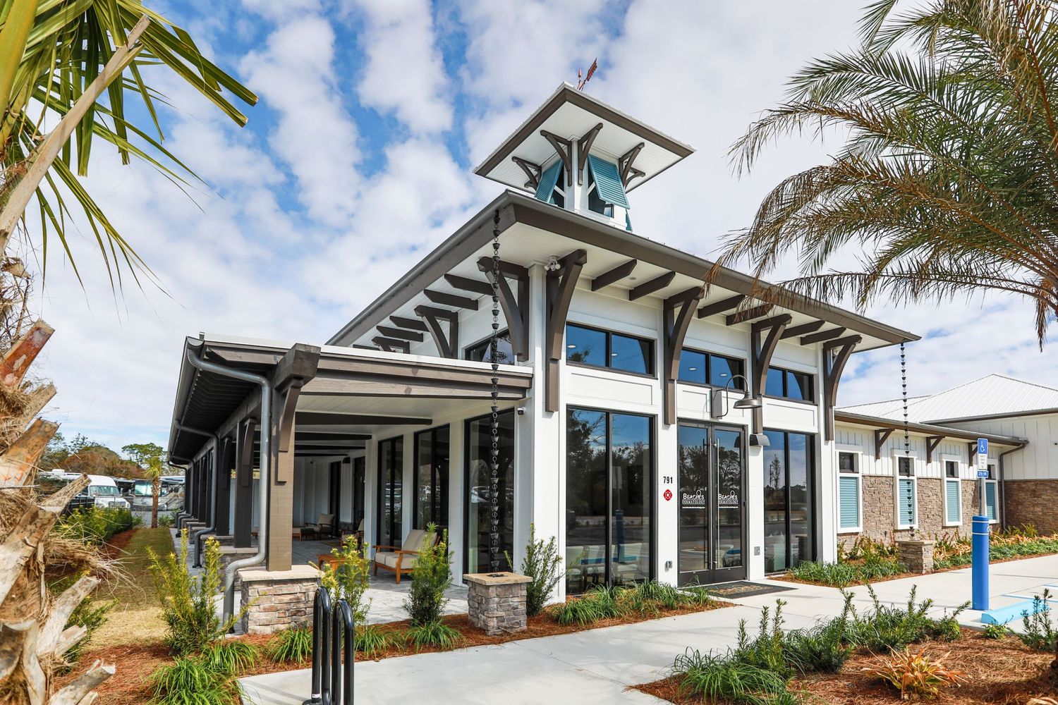 Modern coastal clubhouse building with white trim and large windows surrounded by tropical landscaping and palm trees.