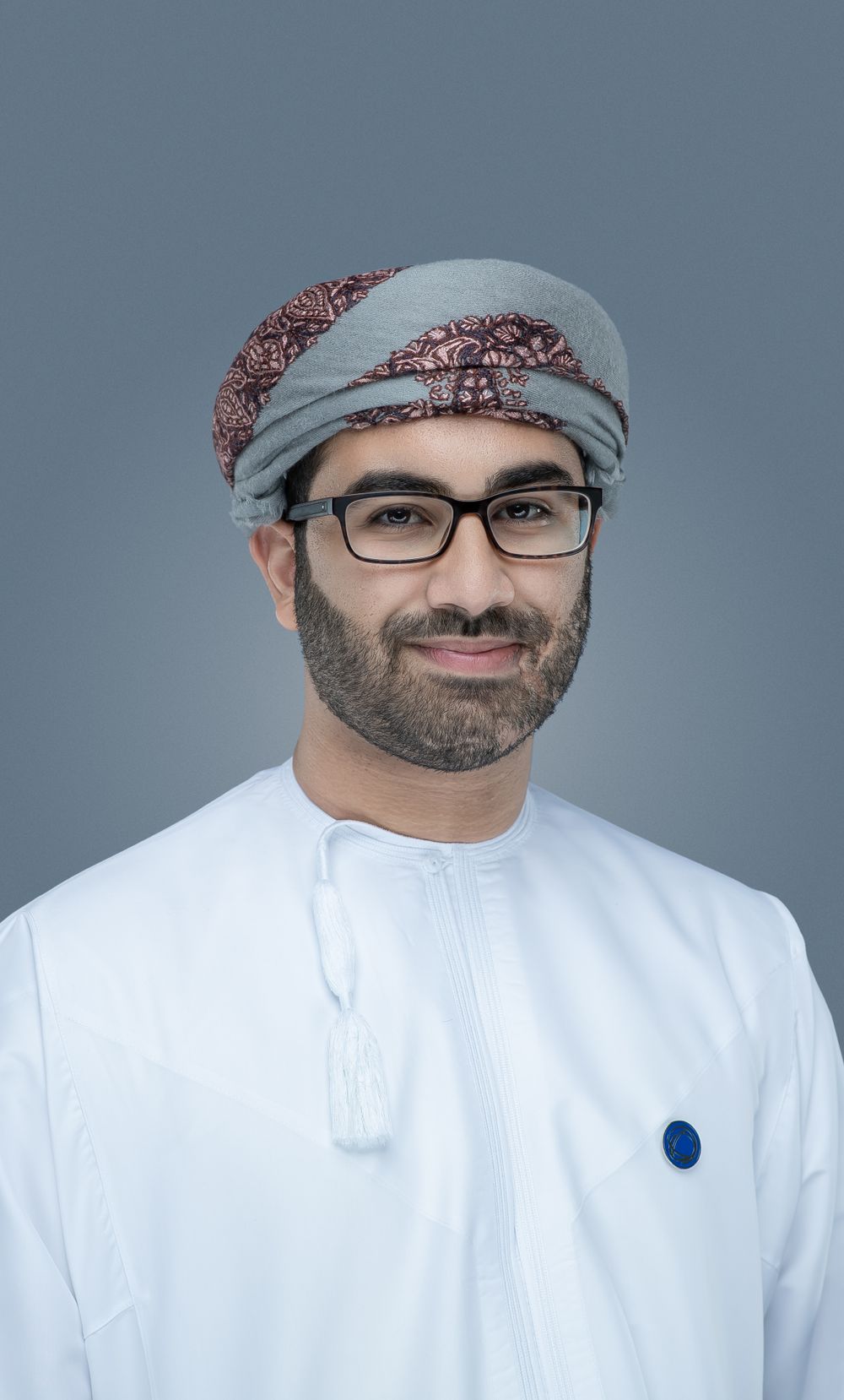 A person wearing traditional Middle Eastern dress including a white thobe and grey patterned headscarf with glasses against grey background.