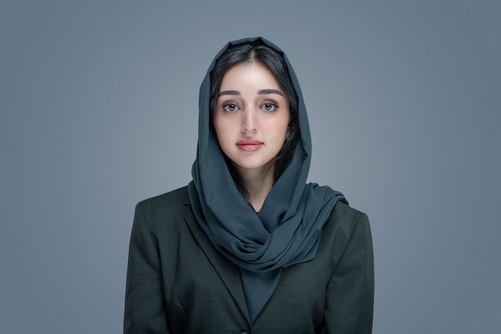 Studio portrait of someone wearing a dark hijab and blazer against a gray background.