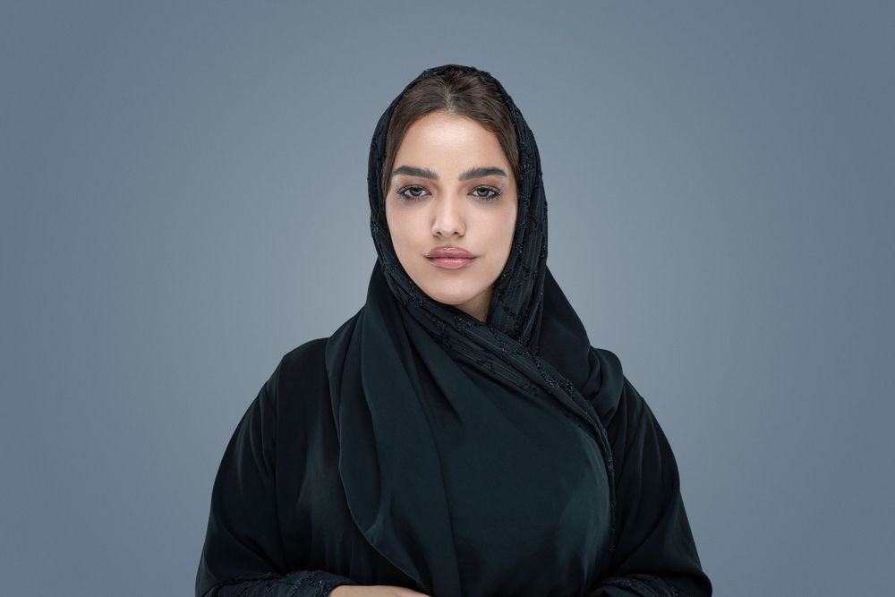 A woman wearing a black hijab poses against a gray studio backdrop for a portrait.
