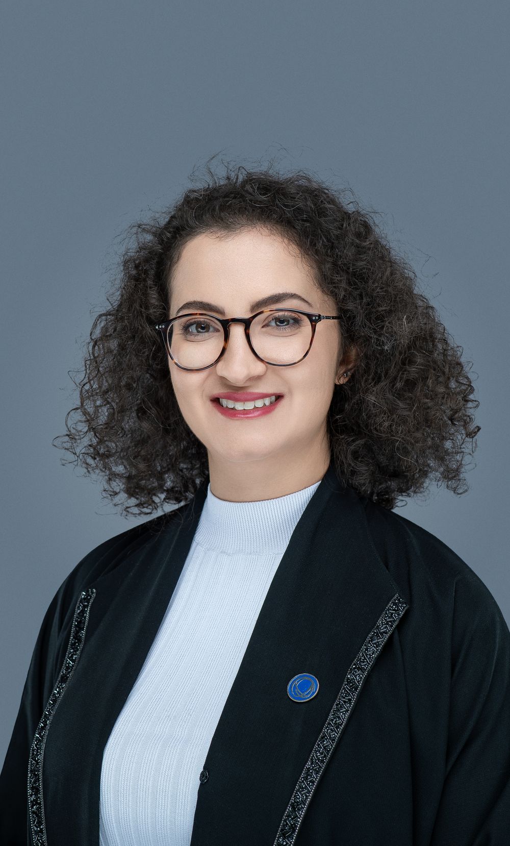 Professional headshot of someone with curly dark hair wearing glasses, a white top and black cardigan against a gray background.
