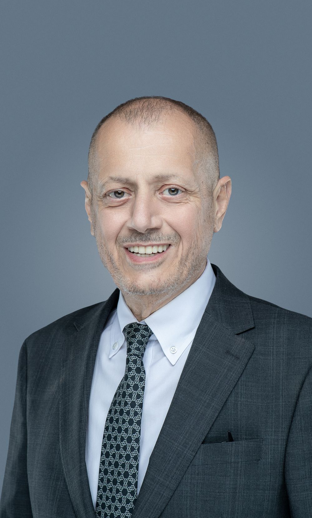 Professional headshot of a smiling business executive wearing a dark suit and patterned tie against a gray background.
