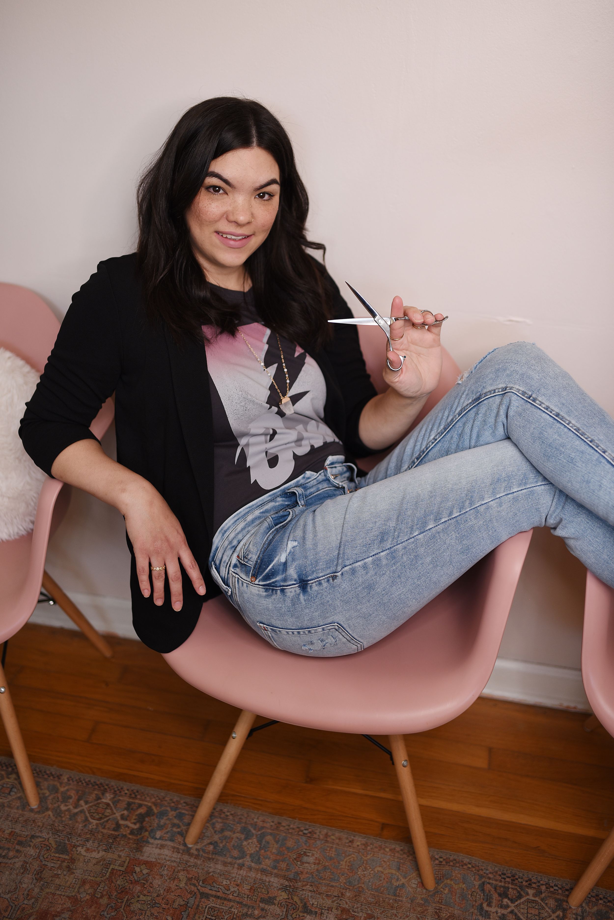 A person relaxes in a pink Eames-style chair wearing jeans, a black cardigan and graphic t-shirt on wooden flooring.