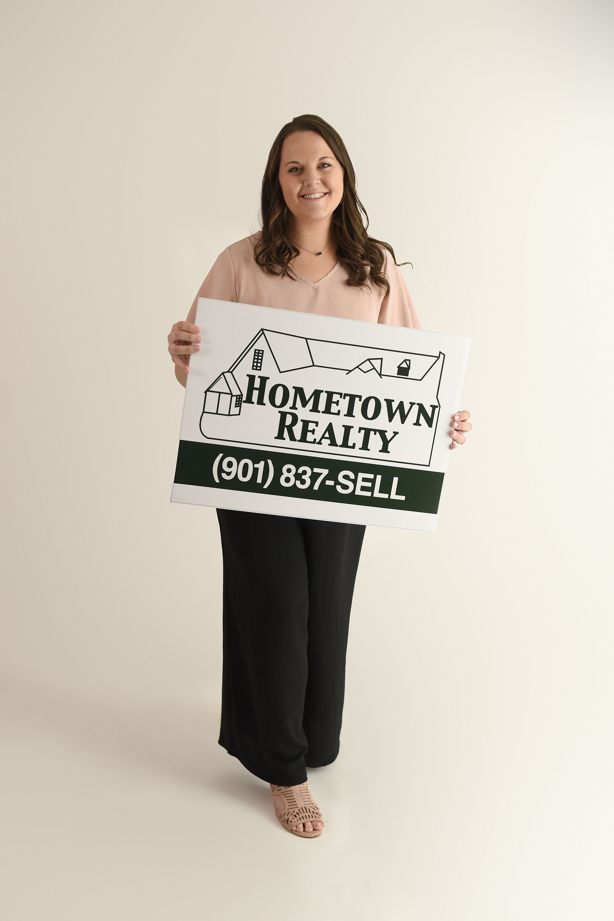 Someone wearing neutral colors stands with a real estate sign showing Hometown Realty and a phone number.