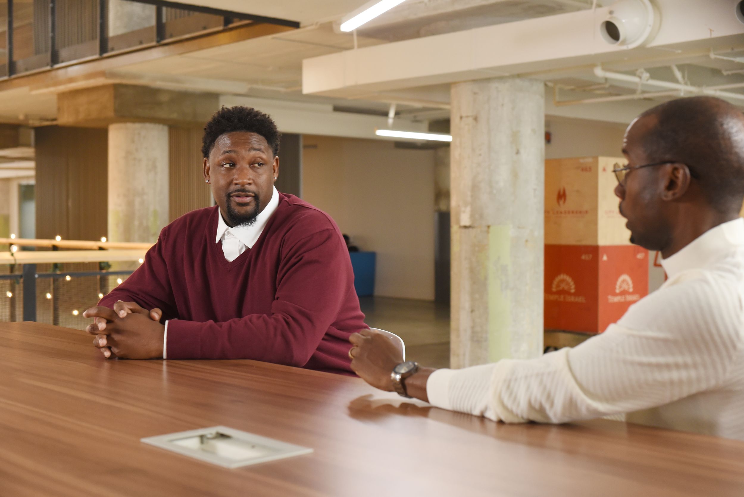 Two colleagues having a discussion while seated at a large wooden conference table in an office space.