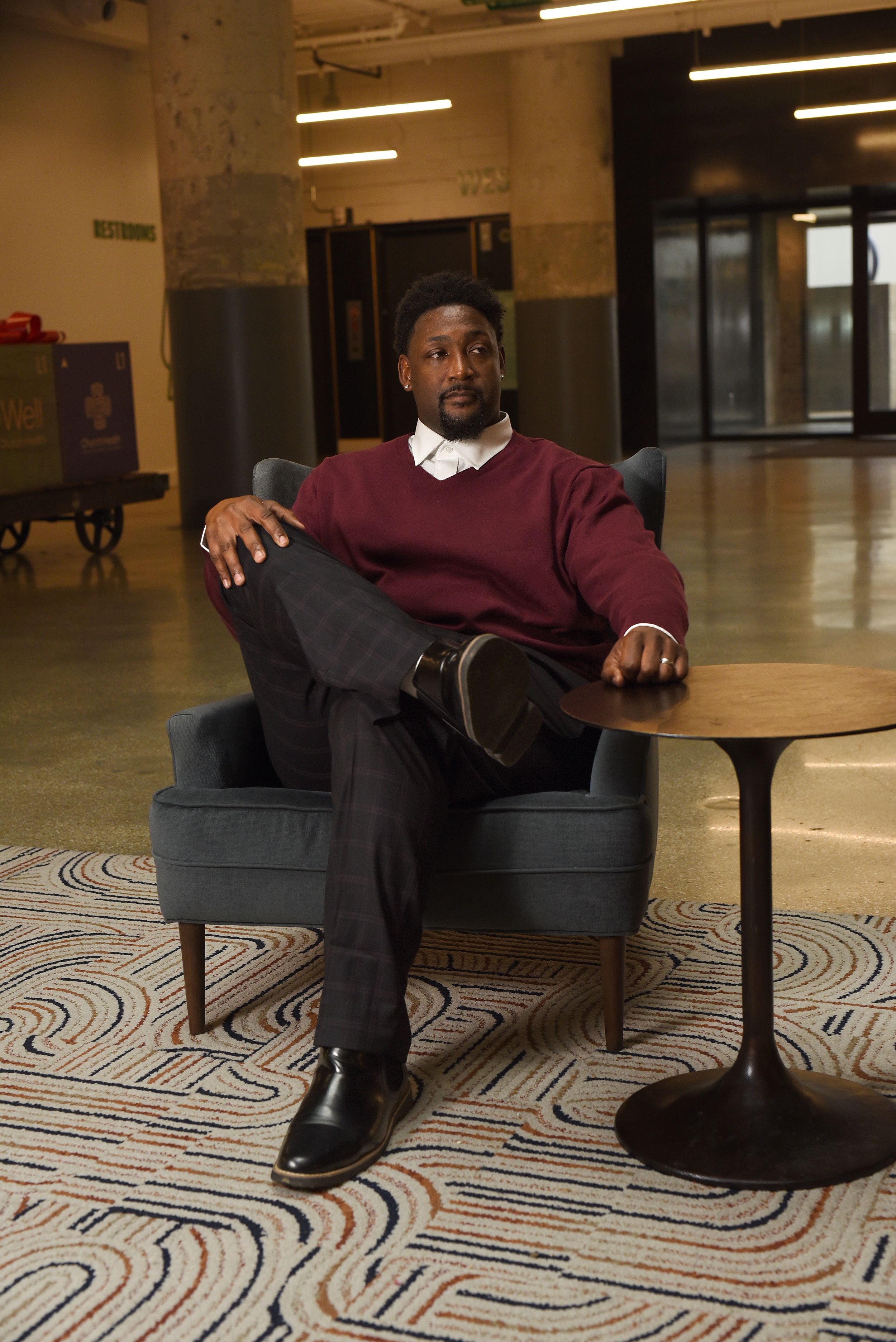 A person sits casually in a gray armchair wearing a burgundy sweater and dark pants in an office-style lobby area.