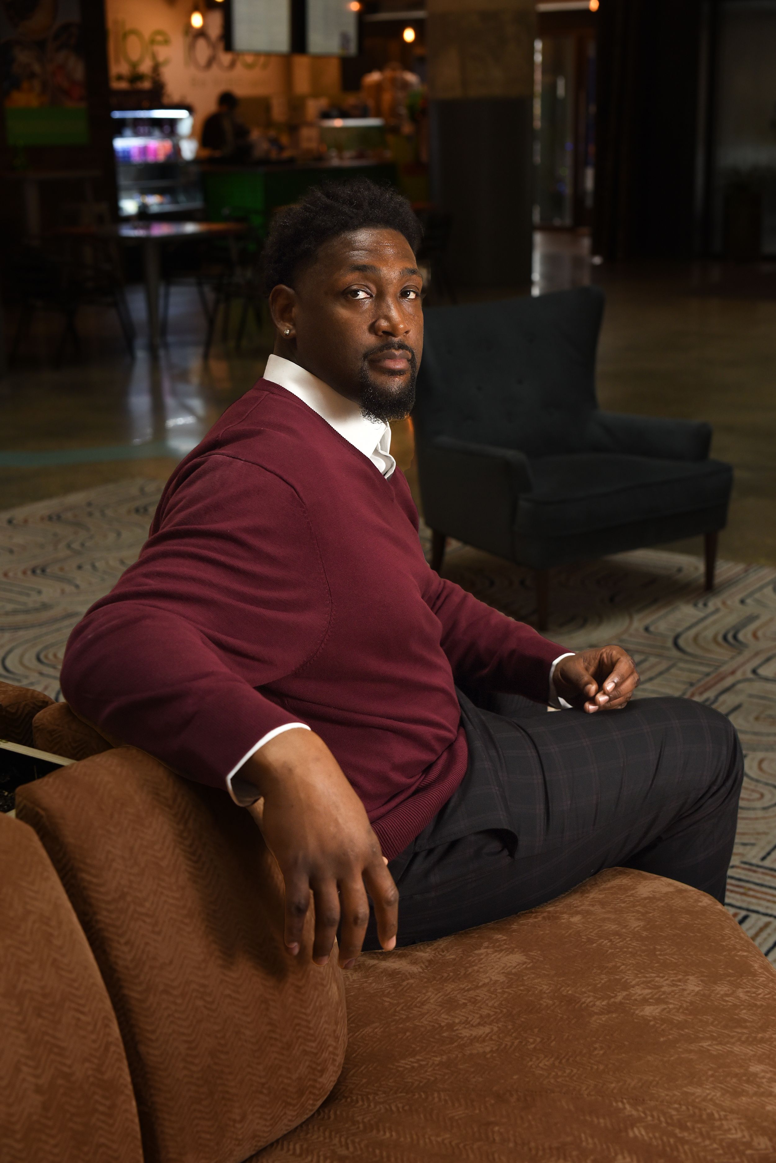 Someone sits casually on a brown couch wearing a burgundy sweater and dress pants in a dimly lit lounge setting.