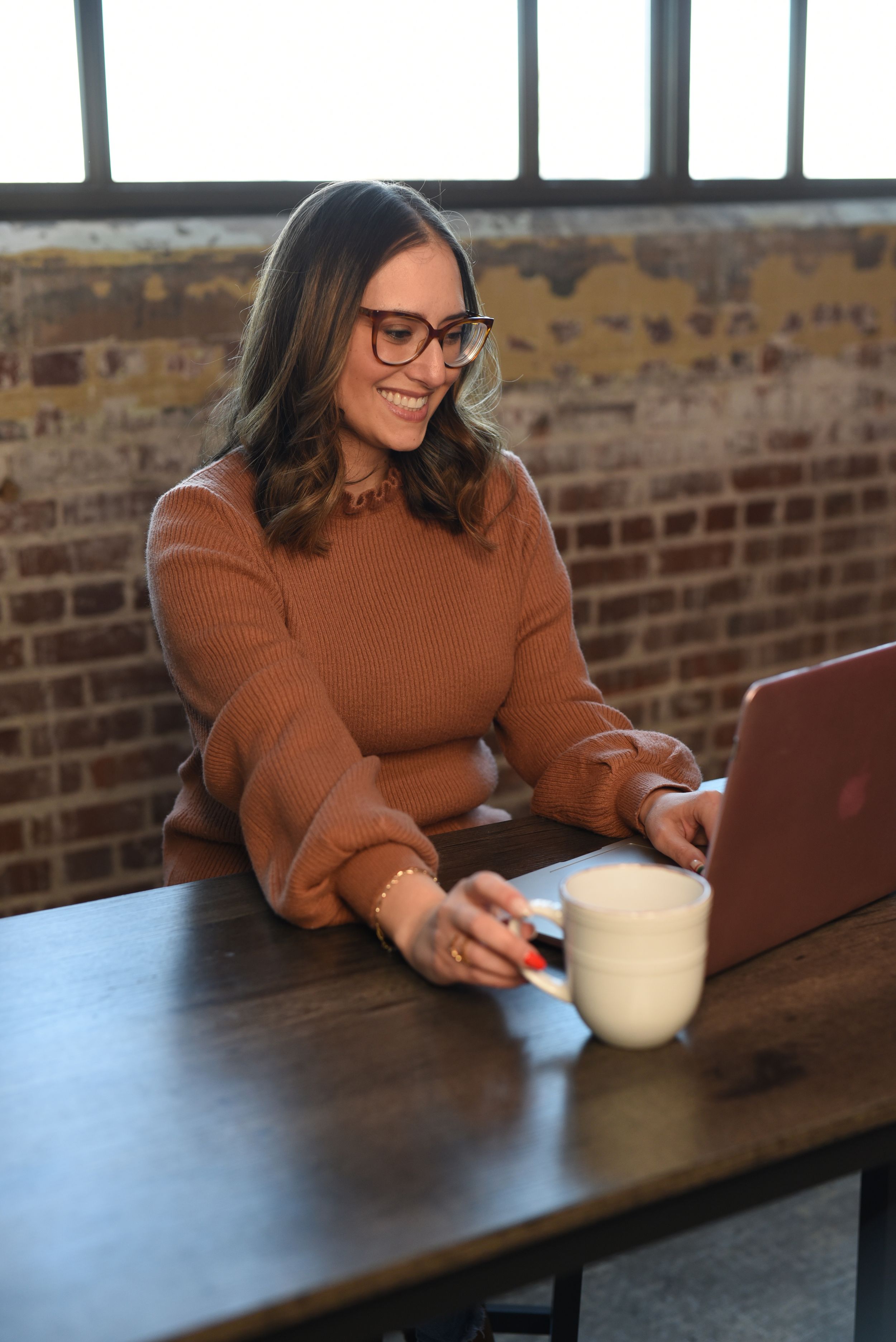 Professional in a brown sweater and glasses working on a laptop at a wooden desk with exposed brick walls behind.