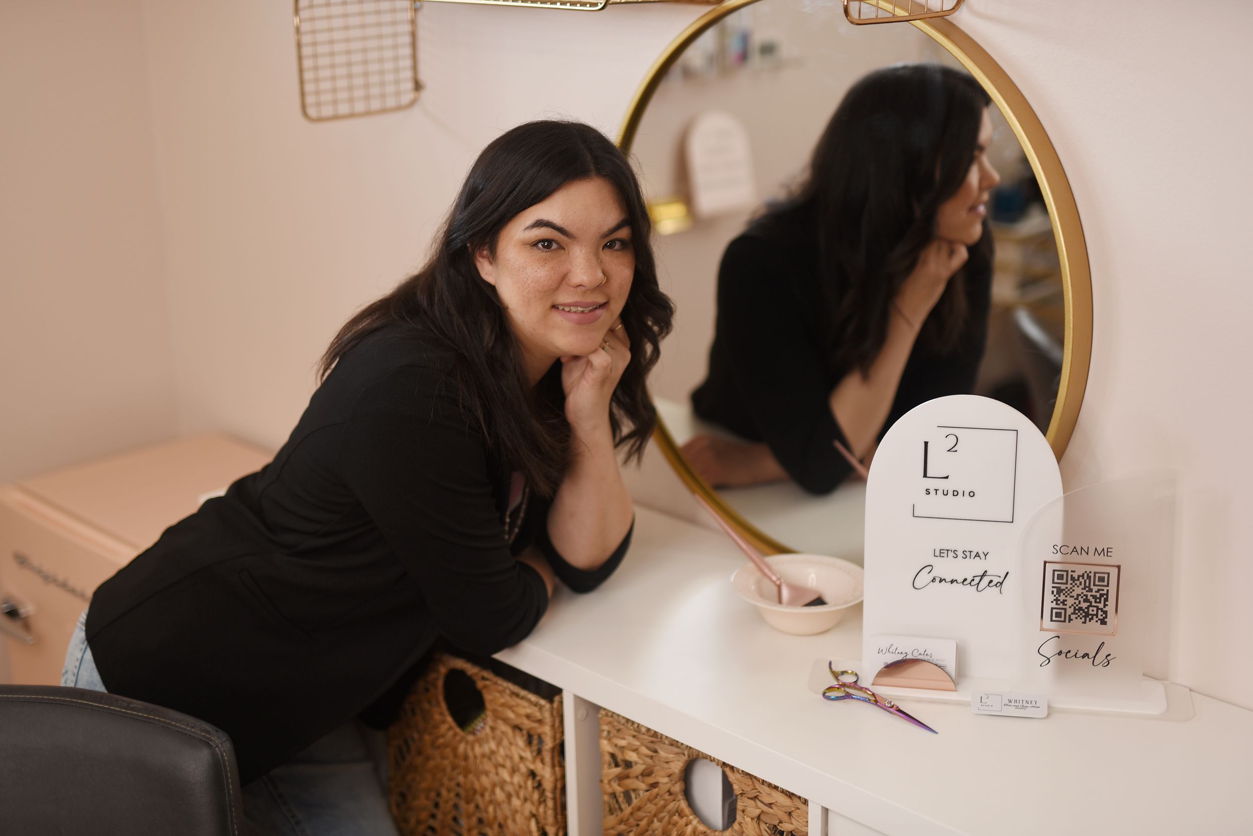 A woman in a black sweater smiles while sitting at a white vanity table with a round mirror.