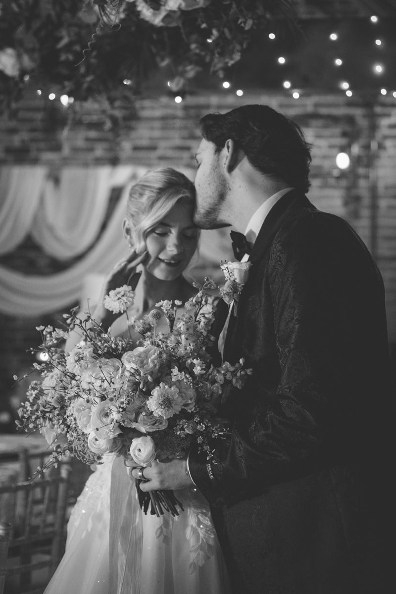 A romantic black and white wedding photo of a couple embracing by a tree in a forest setting with fallen leaves.