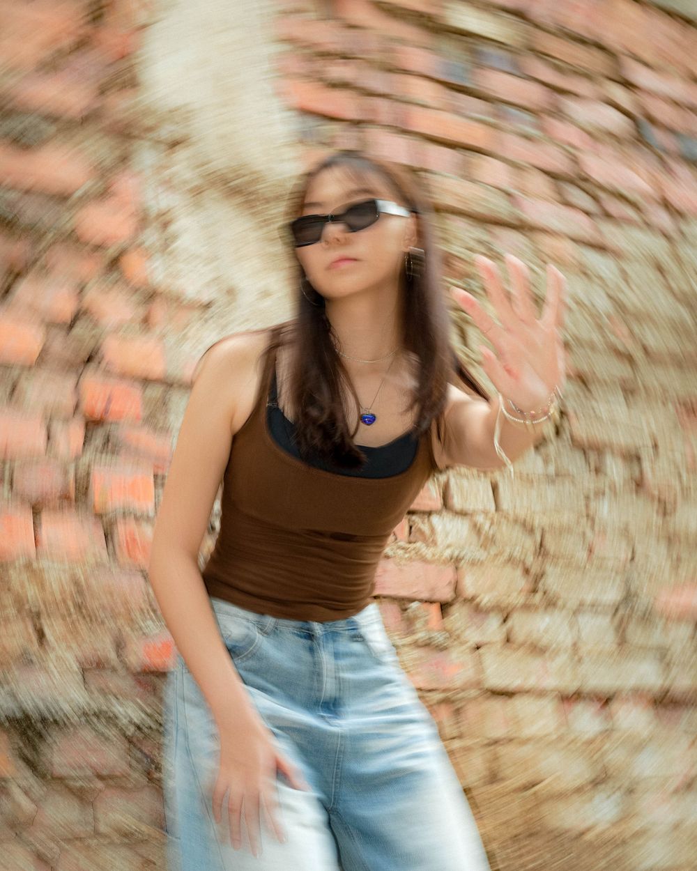 Creative spinning motion blur portrait with brown top and sunglasses against brick wall.