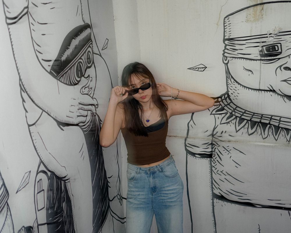 A person in sunglasses poses against a wall with large artistic black and white character drawings.