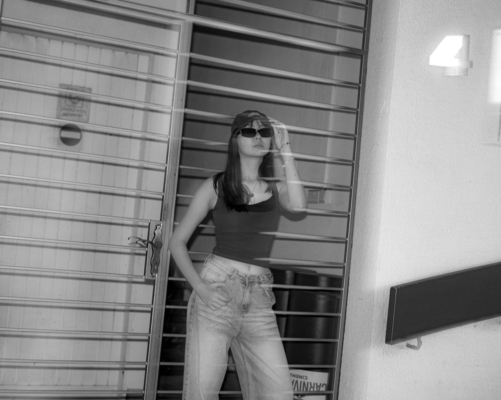 Black and white photo of someone in vintage-style outfit posing against metal grates.