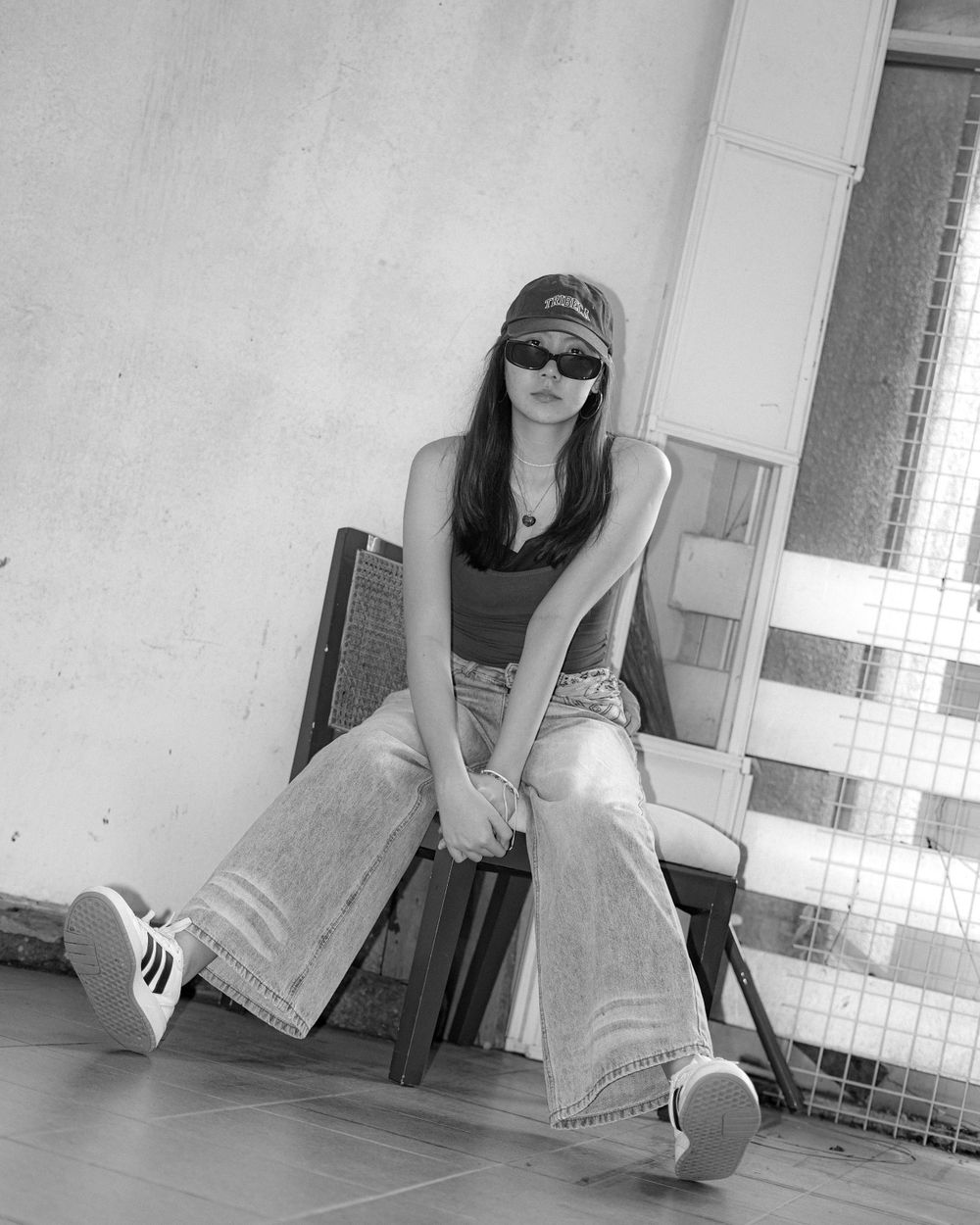 A casual monochrome shot of someone sitting on a wooden chair wearing sunglasses and wide-leg jeans.