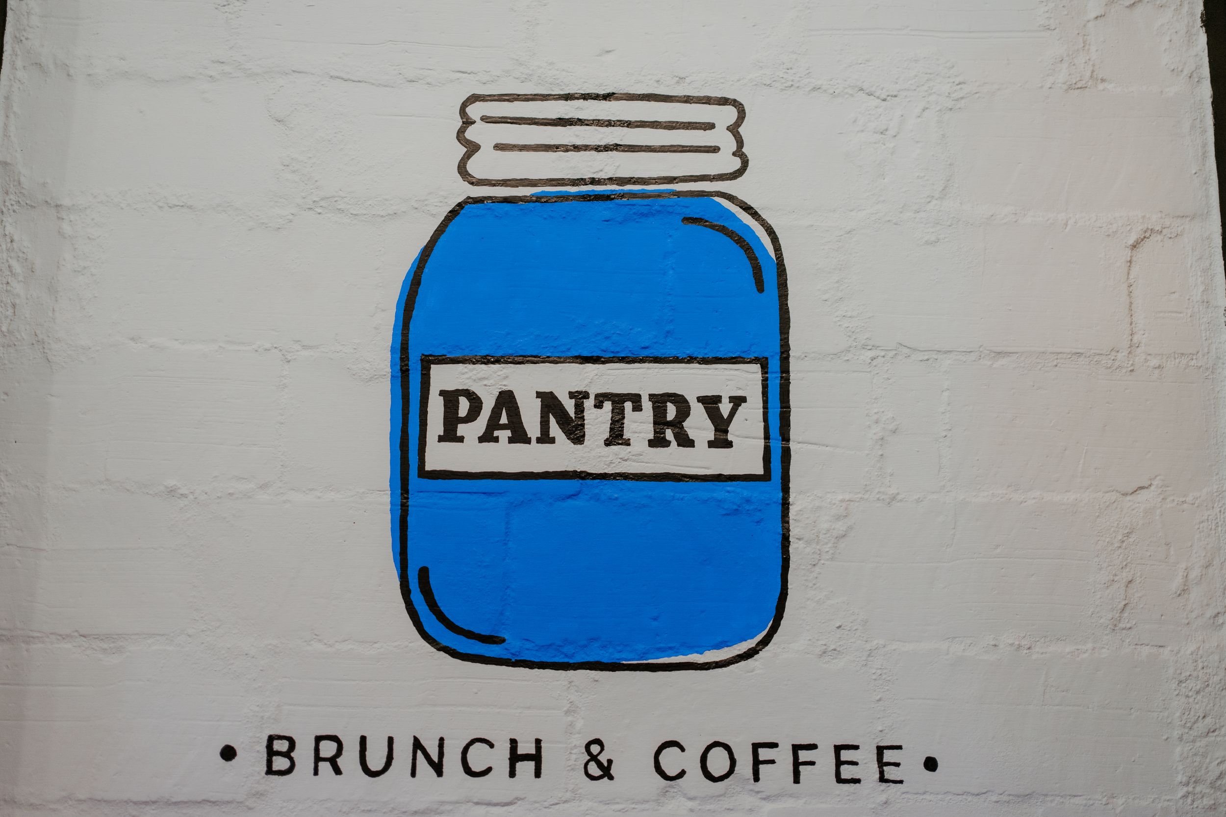 A blue mason jar logo with \"PANTRY\" text and \"BRUNCH & COFFEE\" underneath against a white wall.