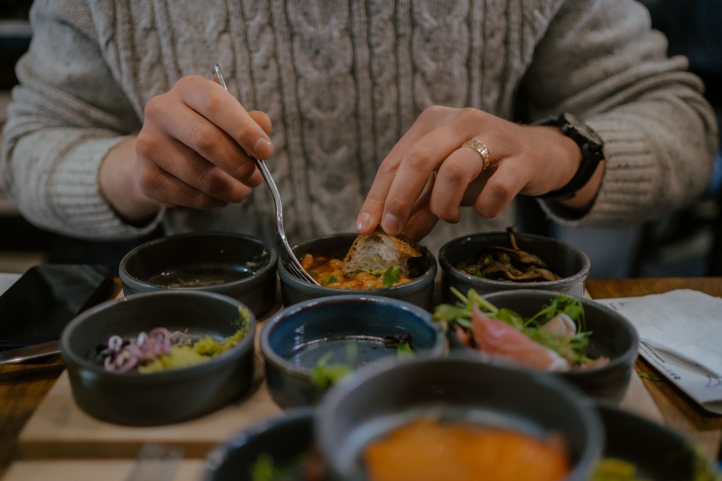 Hands wearing a cable knit sweater serve food from small bowls of colorful sauces and condiments at a restaurant table.