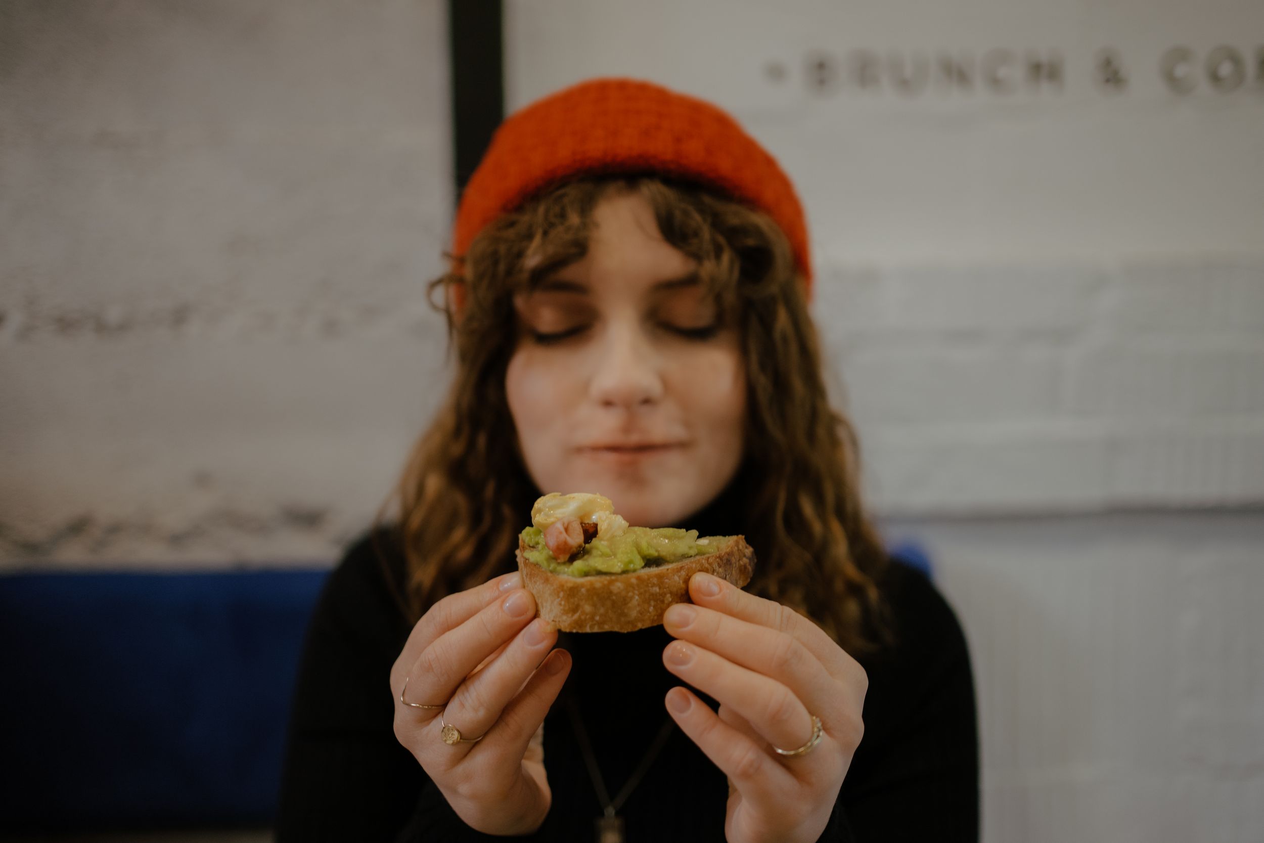 A person in an orange beanie holds and examines a sandwich inside a cafe.