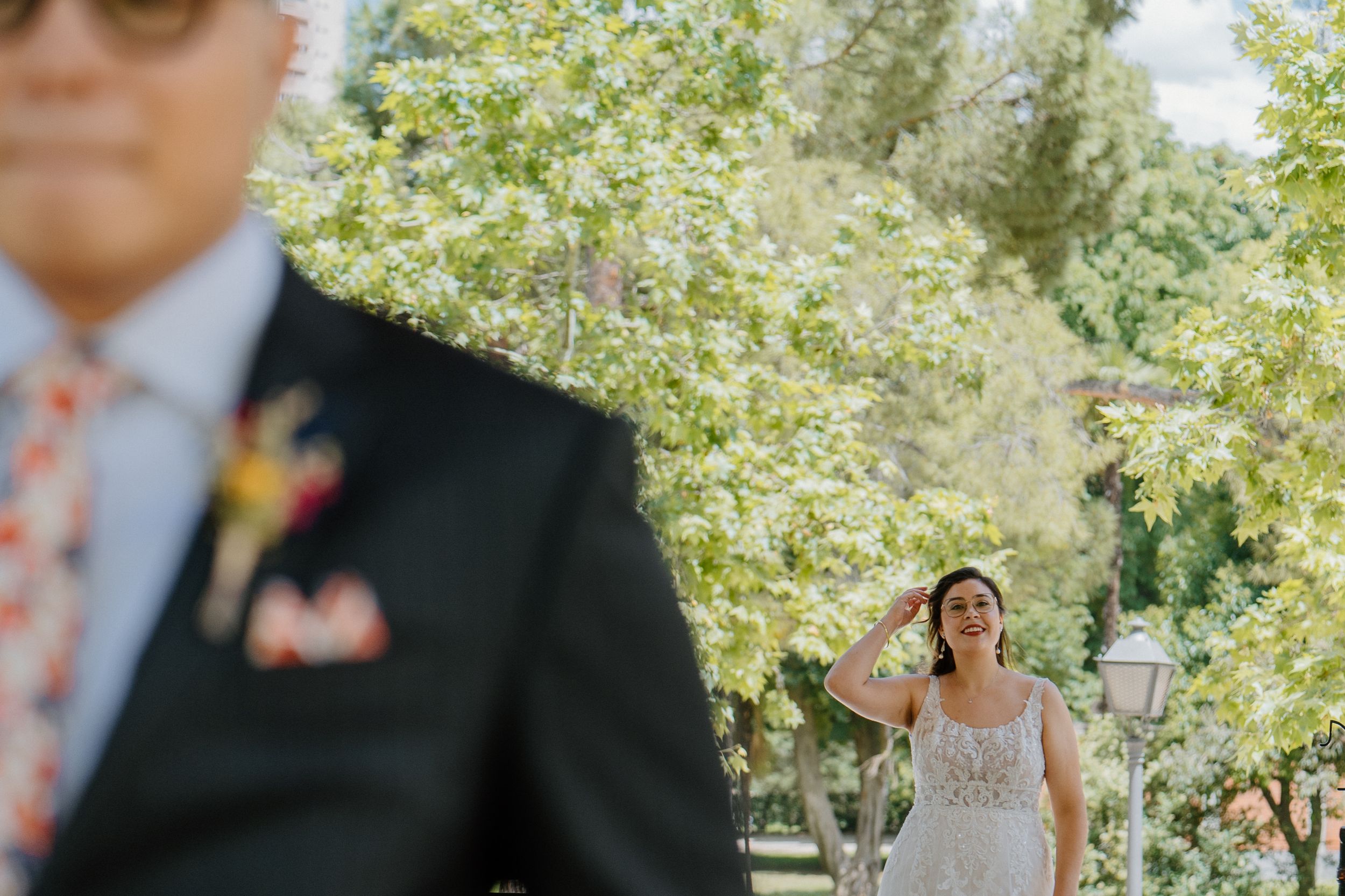 Person in wedding dress approaches someone wearing a black suit and floral tie in a sunny outdoor garden setting.