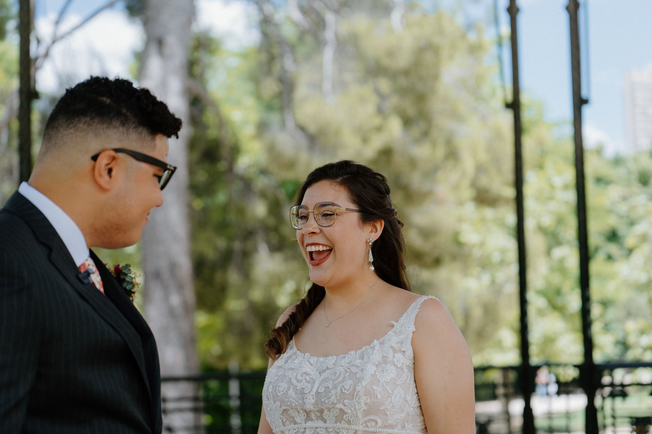 A joyful couple shares a candid moment during their outdoor wedding ceremony beneath tall trees.