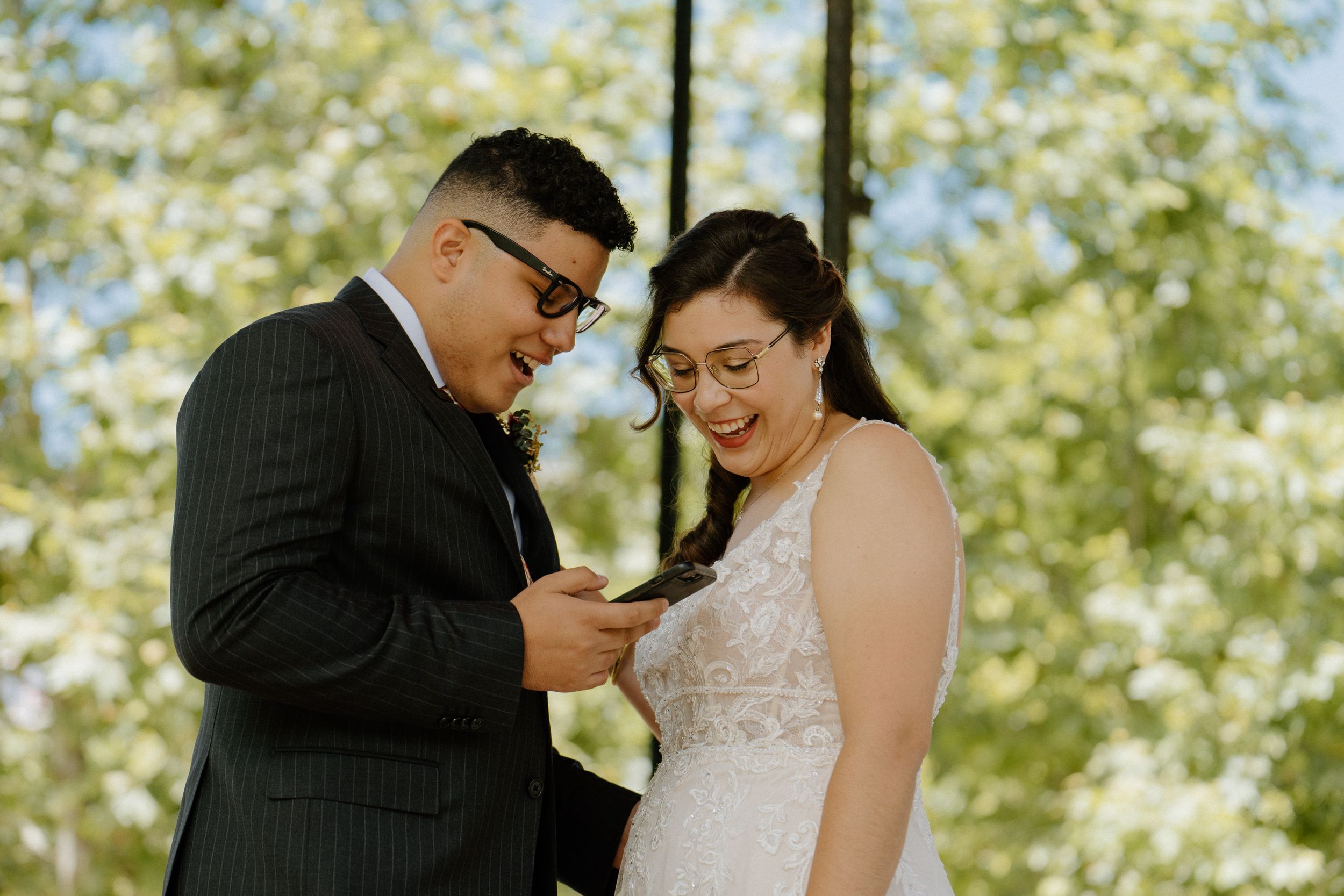 Joyful couple stands together outdoors, looking at phone while wearing formal wedding attire.