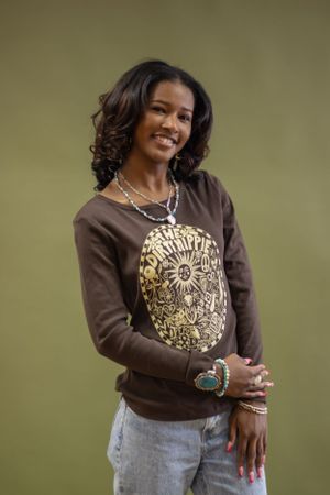 Person wearing a brown sweater with gold embellishments and light jeans poses against a neutral background.