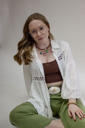 A fashion model wearing a white top top and olive green pants poses on a black stool against a light backdrop during a branding shoot.