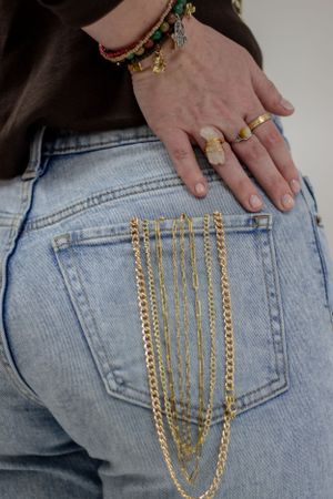 Gold chains hang from back pocket of light wash jeans, with hand wearing multiple rings and bracelets visible.