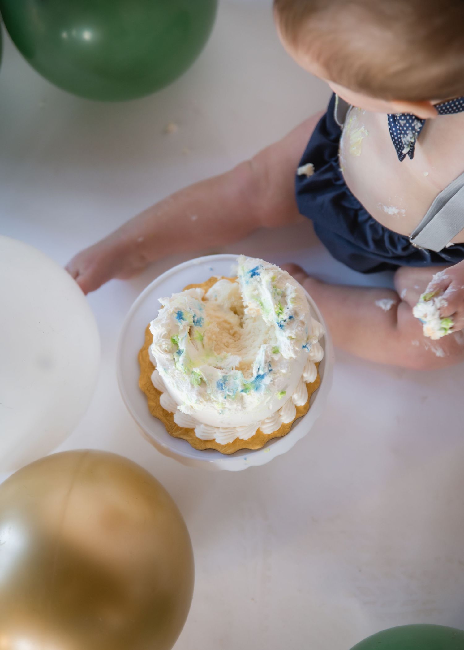 Overhead view of a cupcake with white frosting and sprinkles next to green and gold balloons on a white surface.