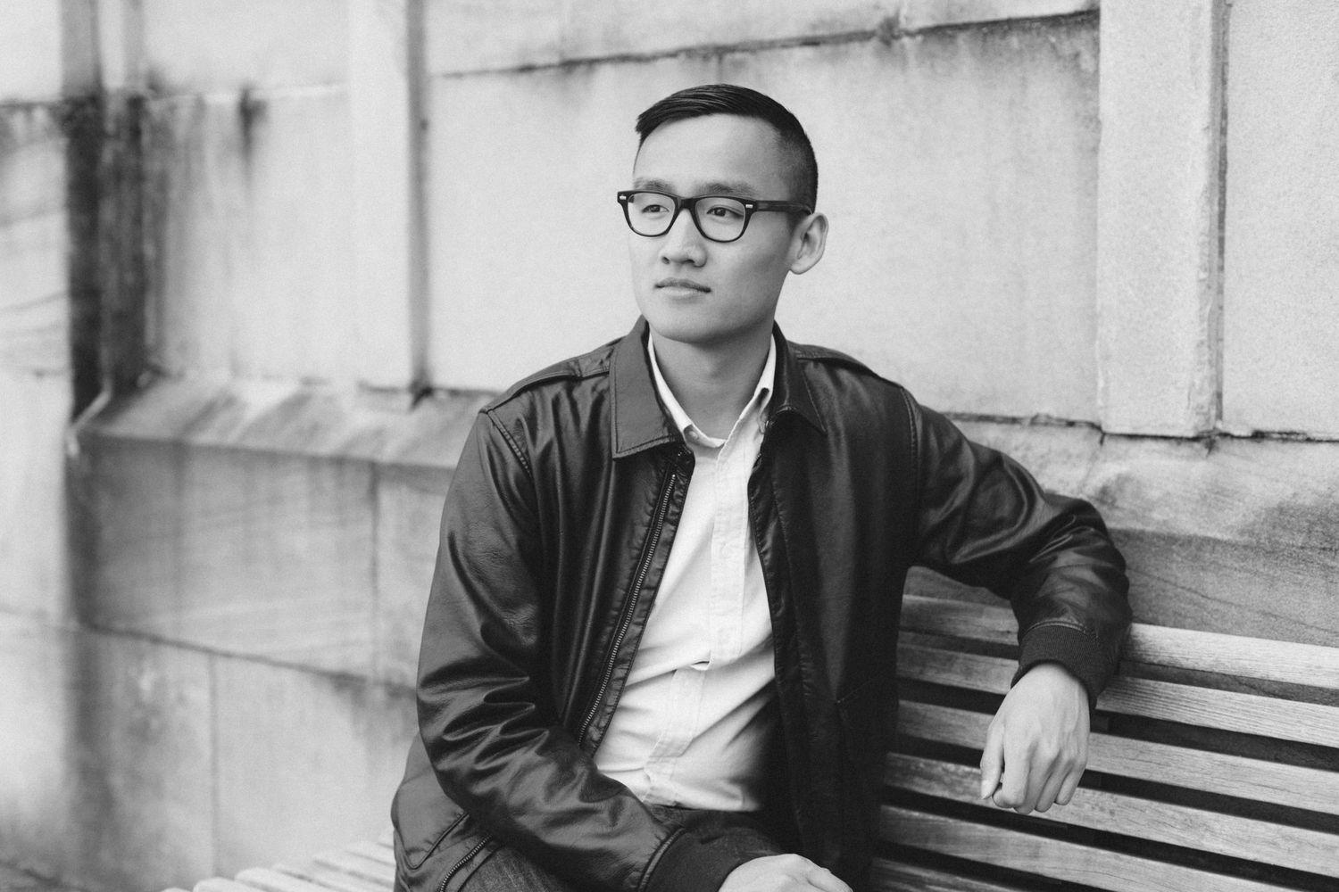 A person in glasses and a leather jacket sitting on a bench in black and white.